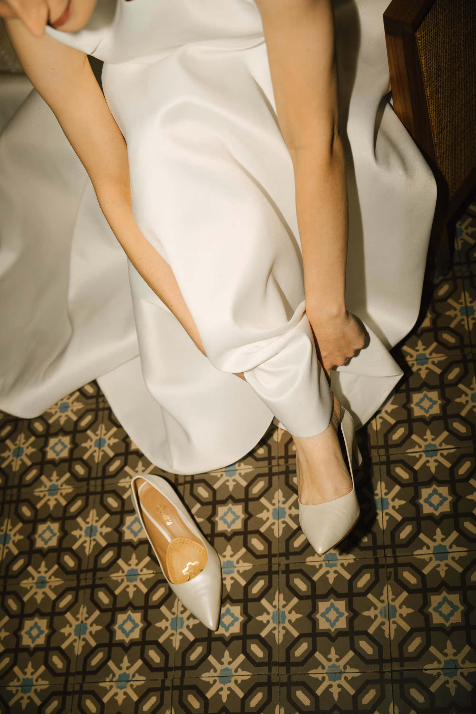 Woman in a white dress putting on one white pointed high heel shoe while the other shoe lies on patterned tile floor.