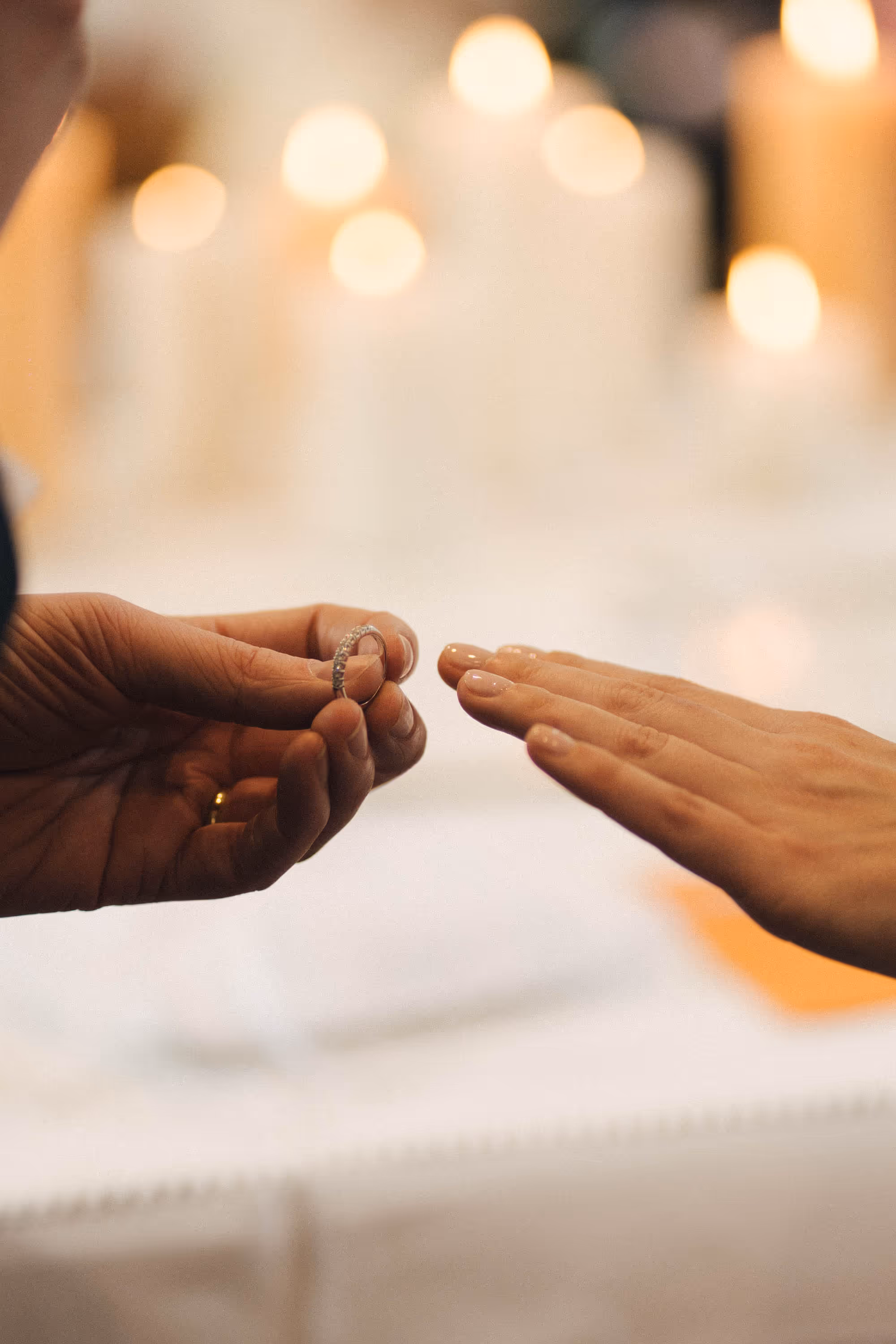 A person holding a diamond-studded ring near another person's outstretched hand against a background of blurred candle lights.