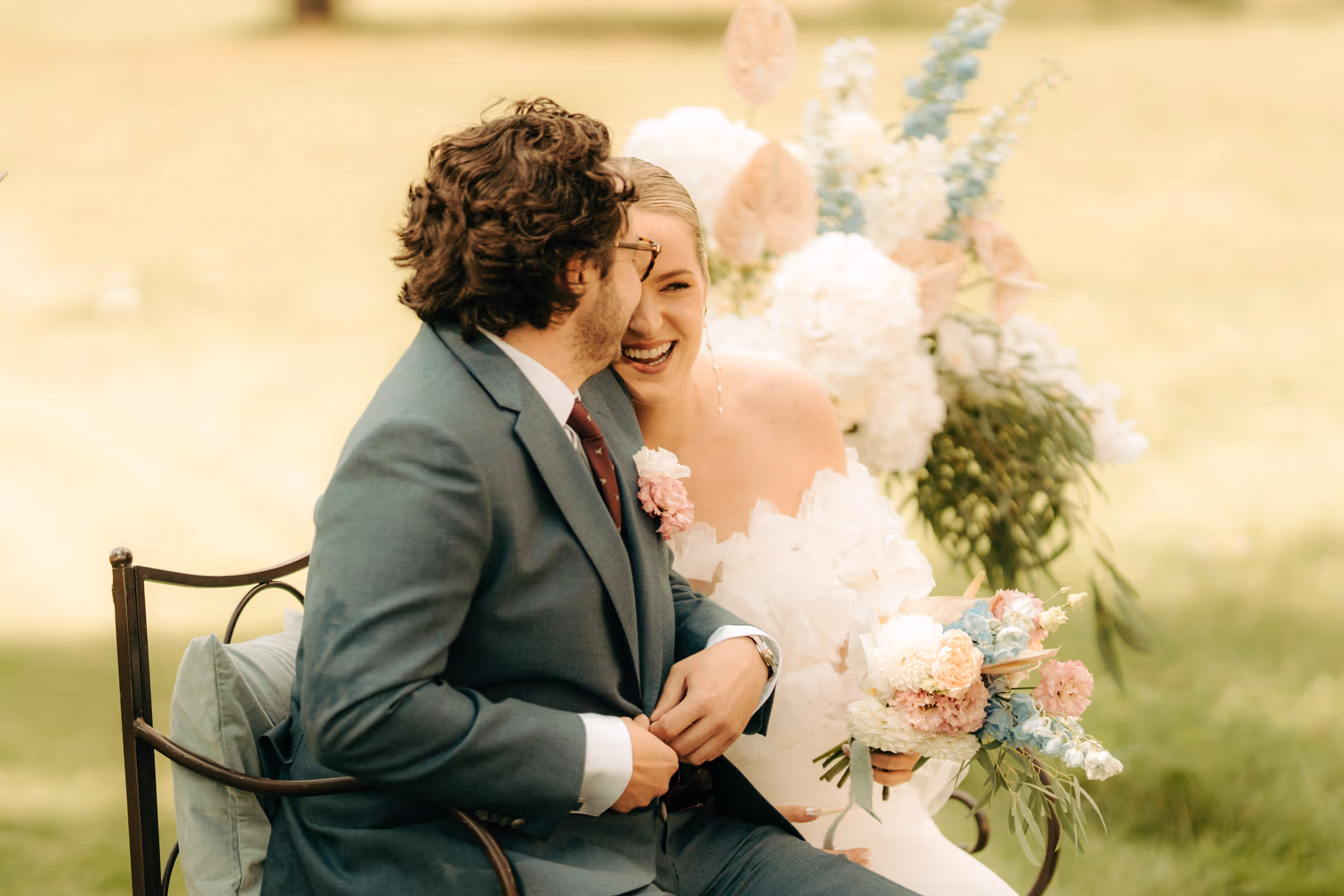 Bride and groom sitting on a bench outdoors, smiling and holding a bouquet of pastel flowers.