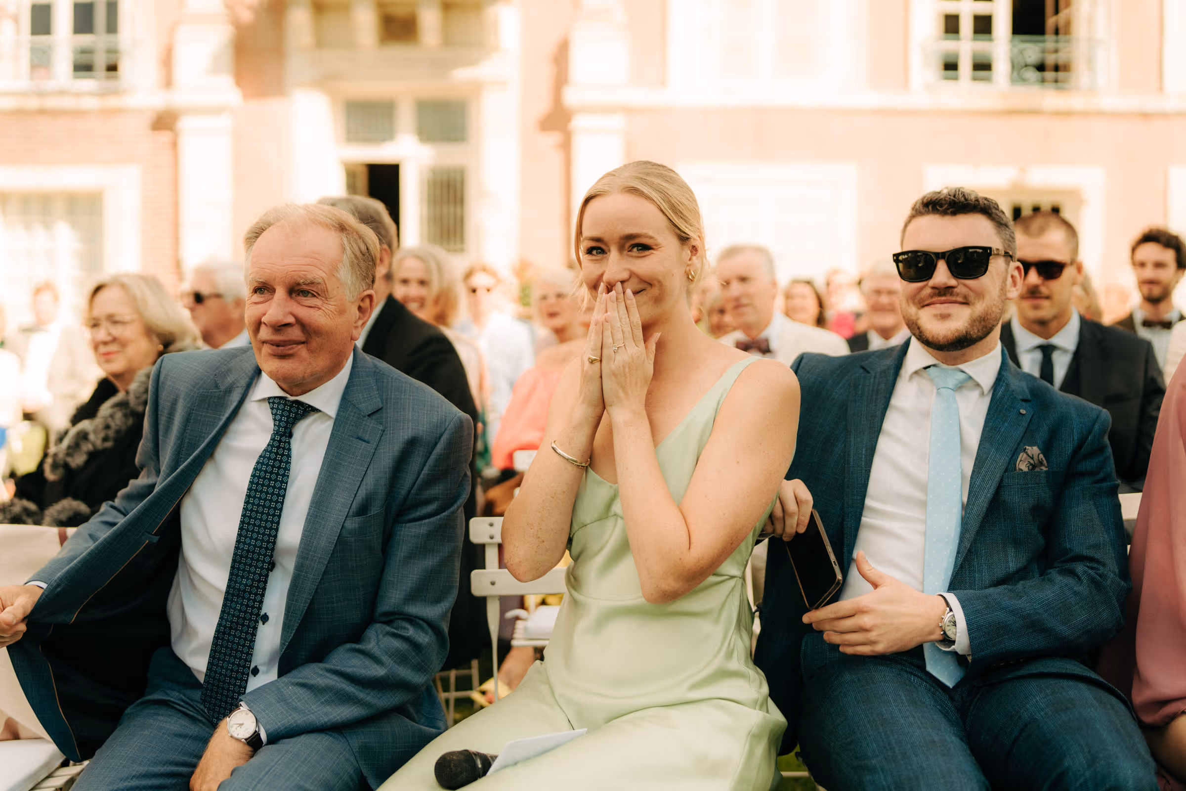Three people seated outdoors at a formal event, with a woman in a light green dress smiling and covering her mouth between two men in suits.
