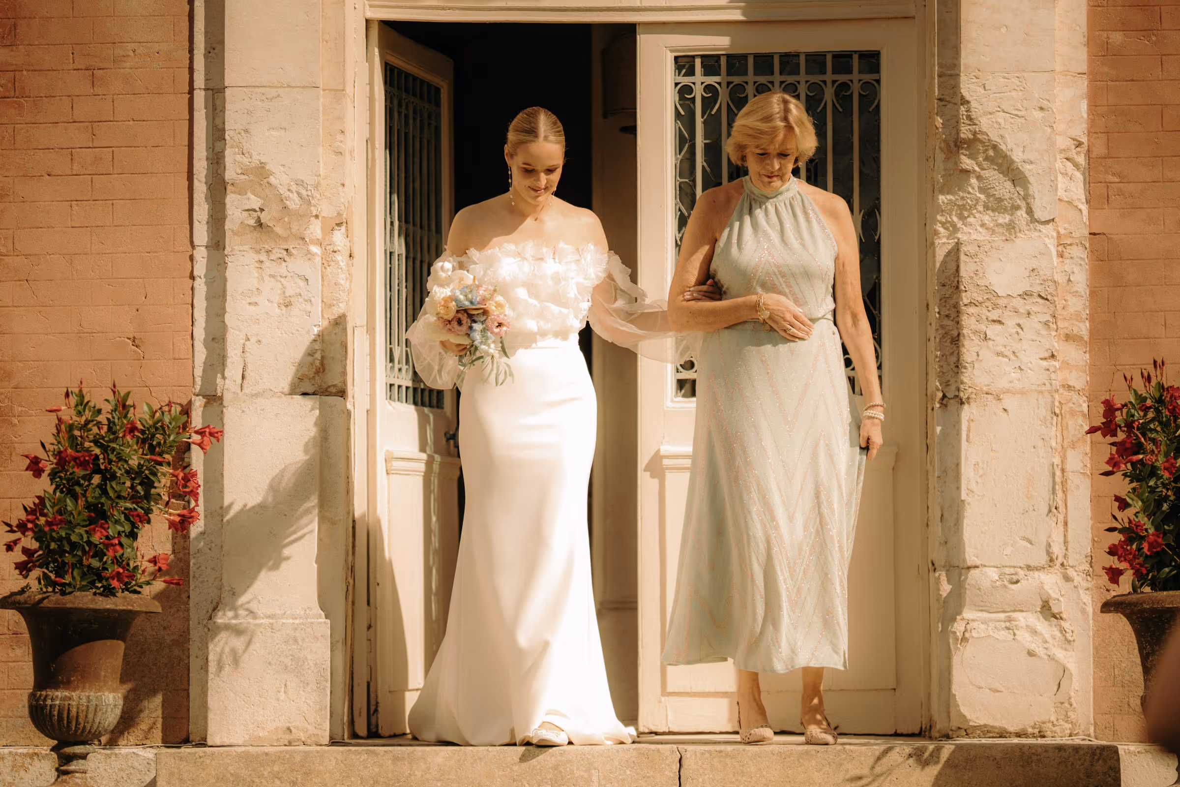 Bride in white dress holding a bouquet, walking arm in arm with an older woman in a light green dress outside a stone building.
