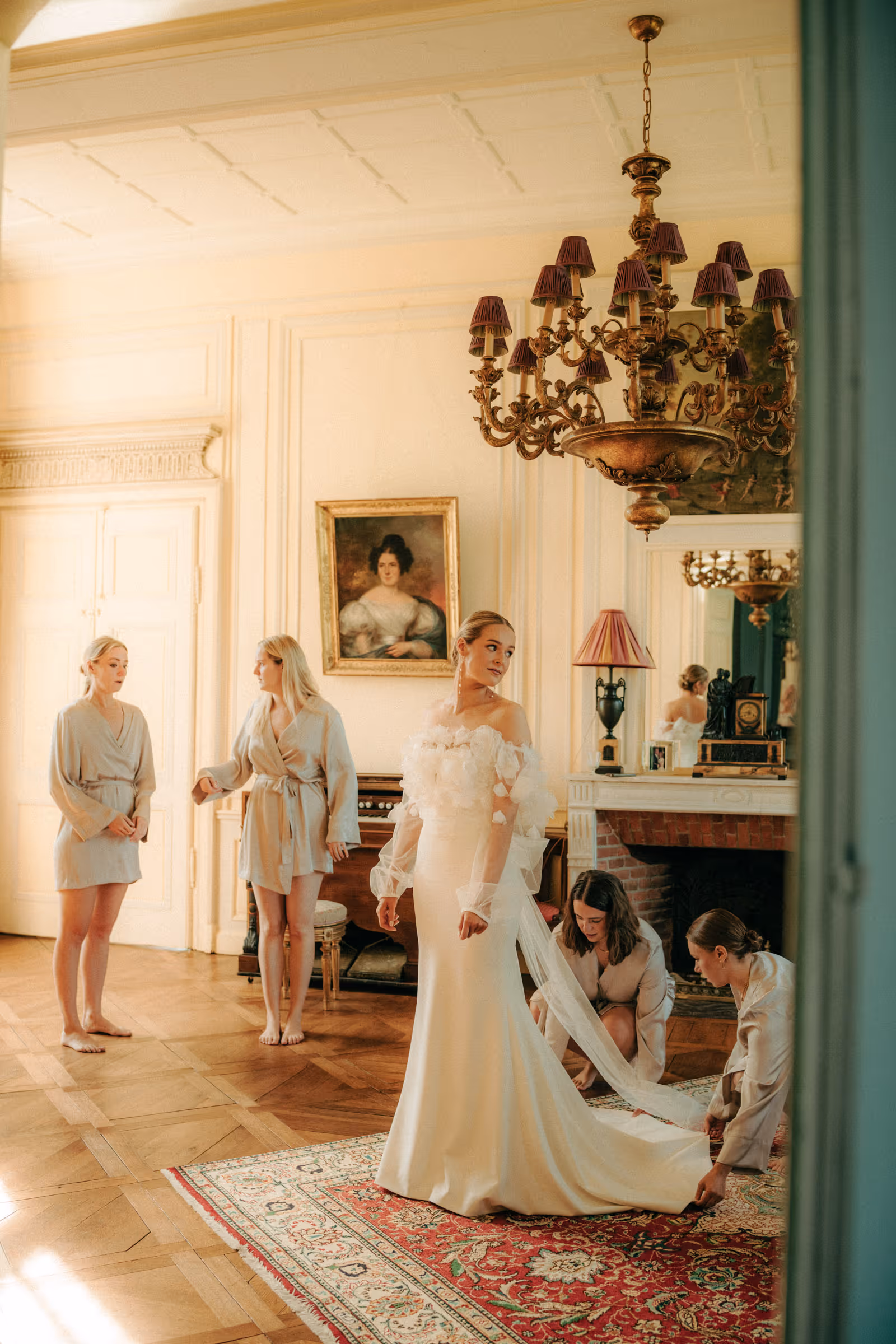 Bride in an off-shoulder white wedding gown stands on a patterned rug in a vintage room as two women adjust her dress and two others in robes stand nearby.