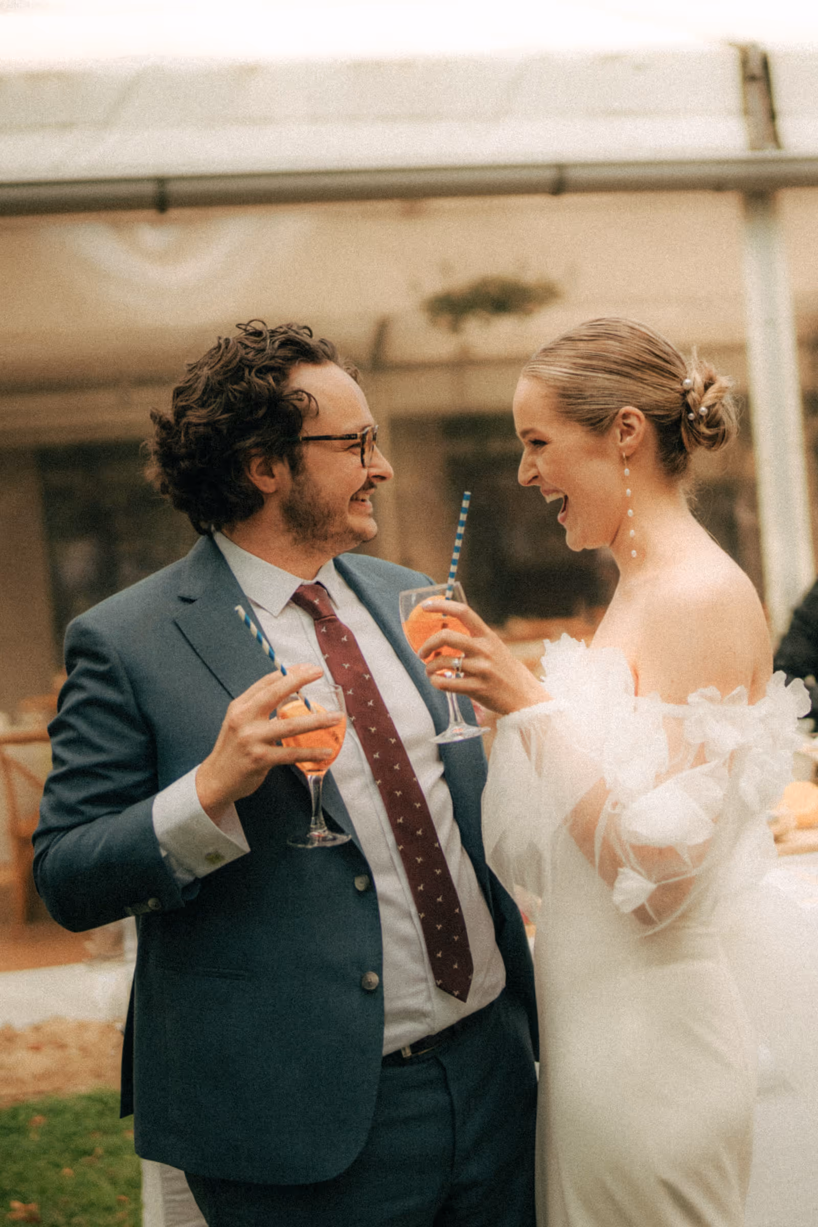 Man in suit and woman in white dress laughing and holding drinks with orange beverage and blue striped straws.