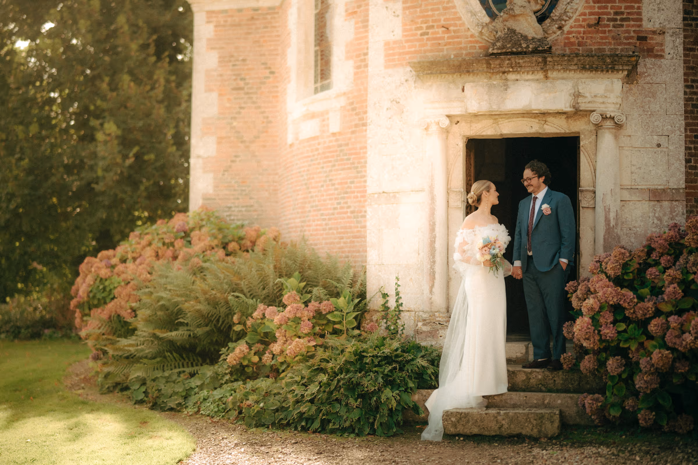 Bride and groom holding hands and smiling at each other outside a stone church doorway surrounded by flowering bushes.