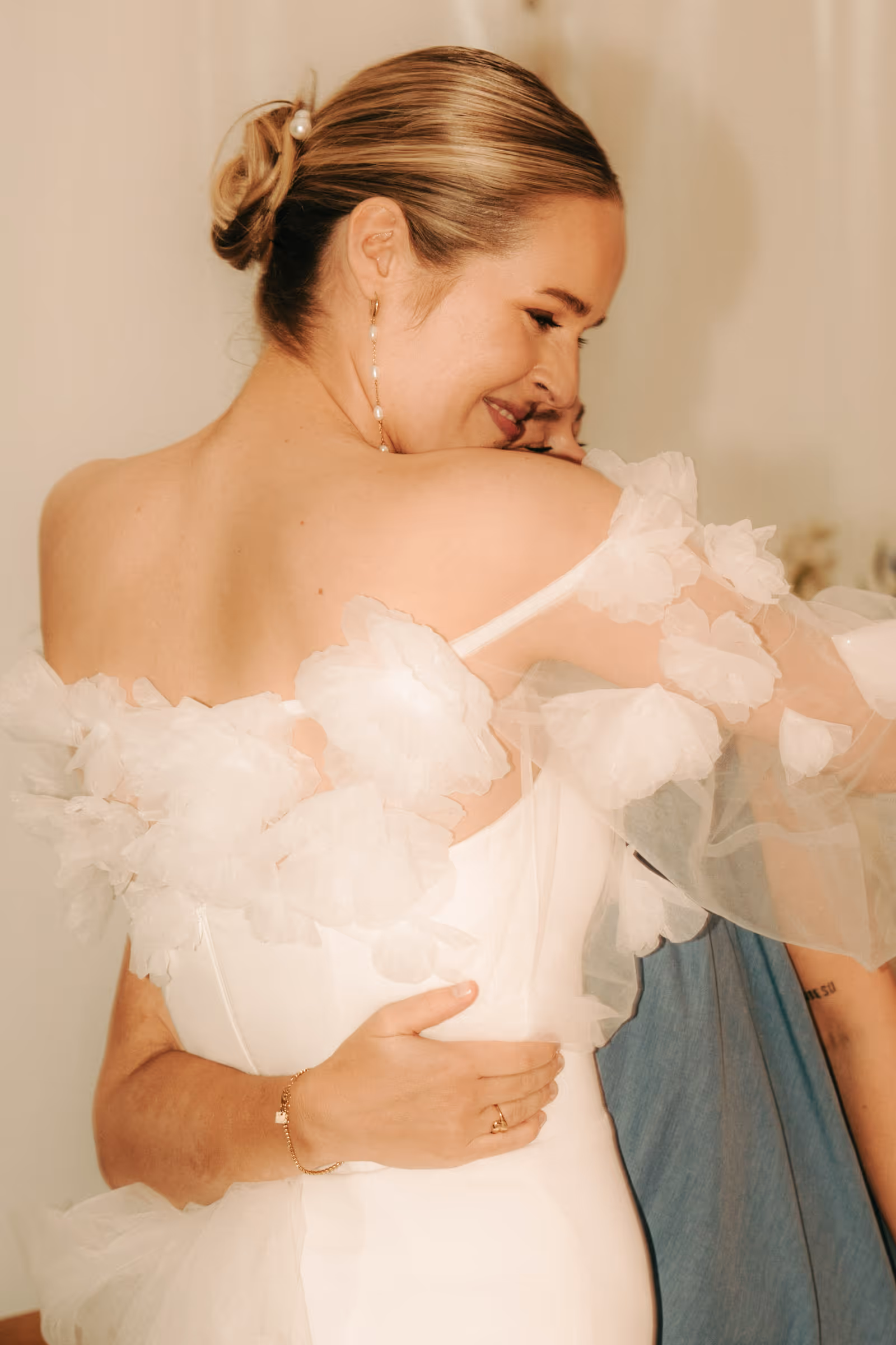 Woman in an off-shoulder white dress with floral tulle hugging another person in a blue outfit.