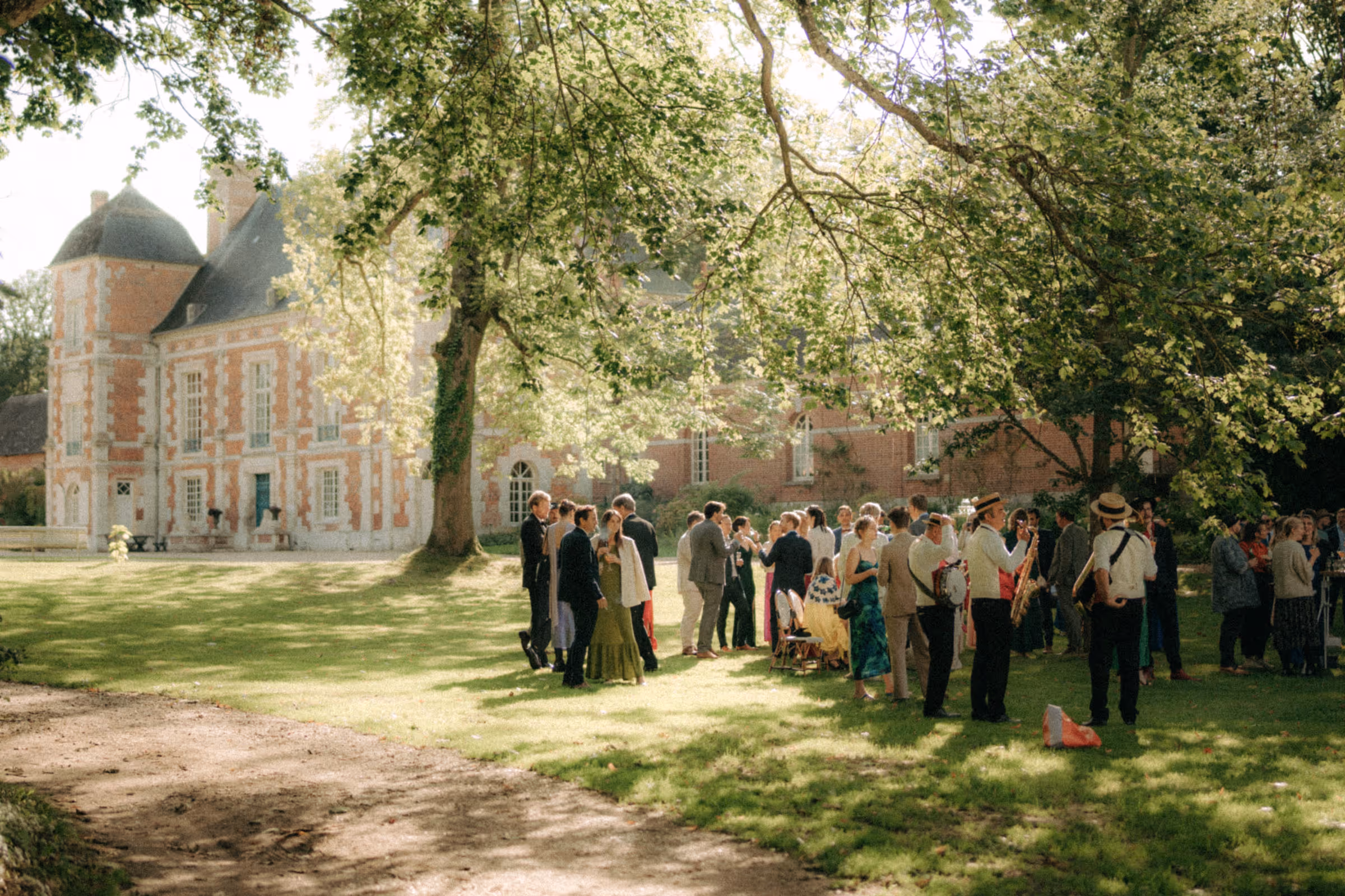 Group of people socializing on sunlit grass under large trees near a historic brick building.