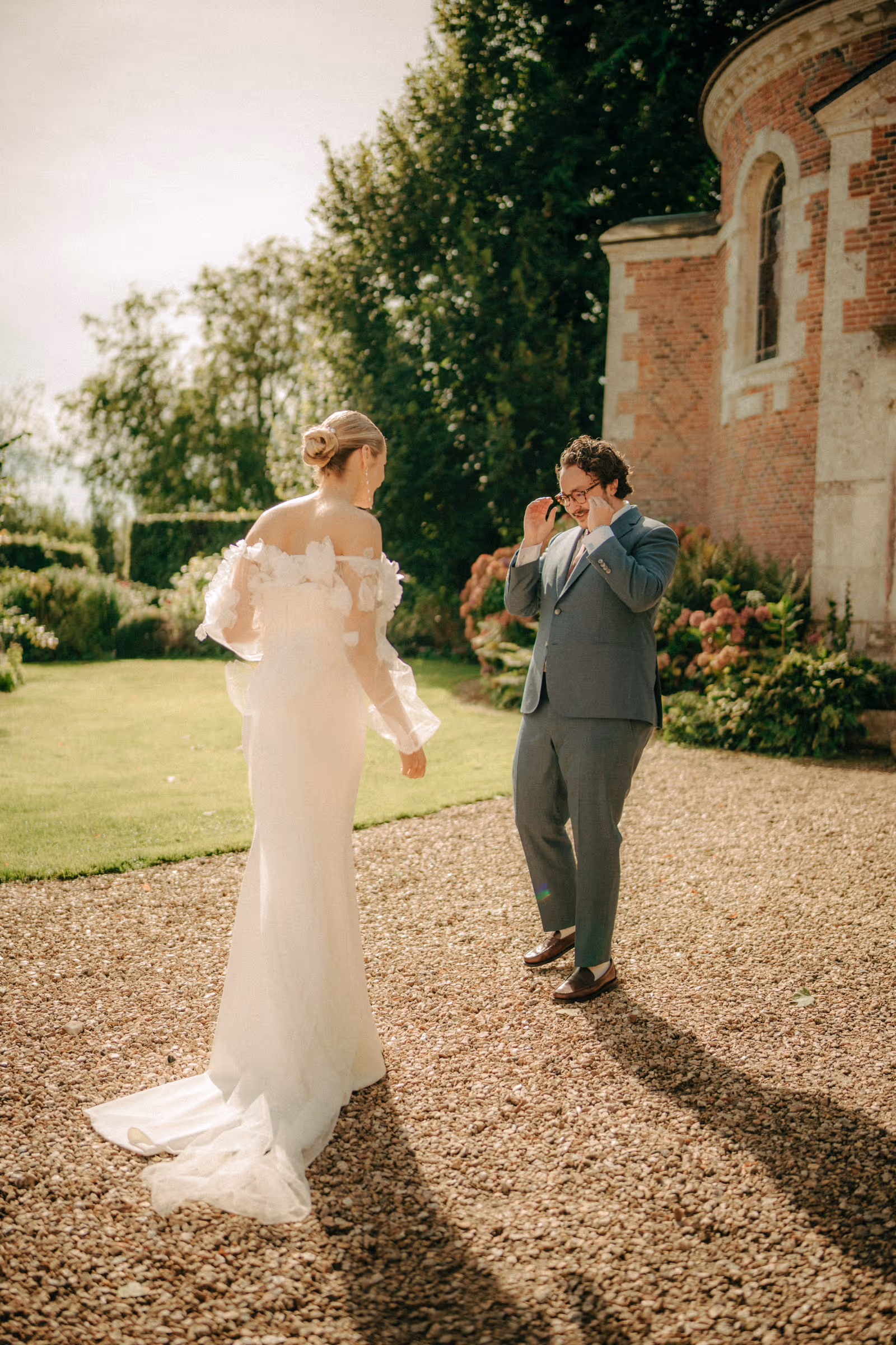 Bride in a white wedding dress approaching a groom in a blue suit who is adjusting his glasses in an outdoor garden setting.
