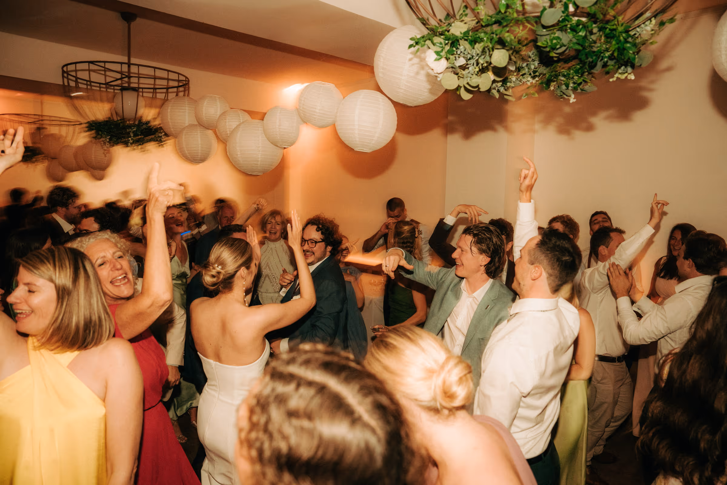 Group of people dancing and celebrating indoors under hanging paper lanterns and greenery decorations.