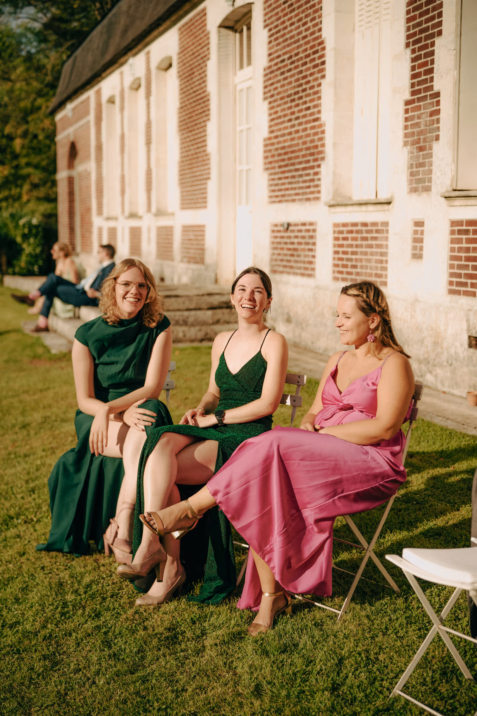 Three women in elegant green and pink dresses sitting on chairs on grass outside a brick building, smiling and enjoying sunny weather.