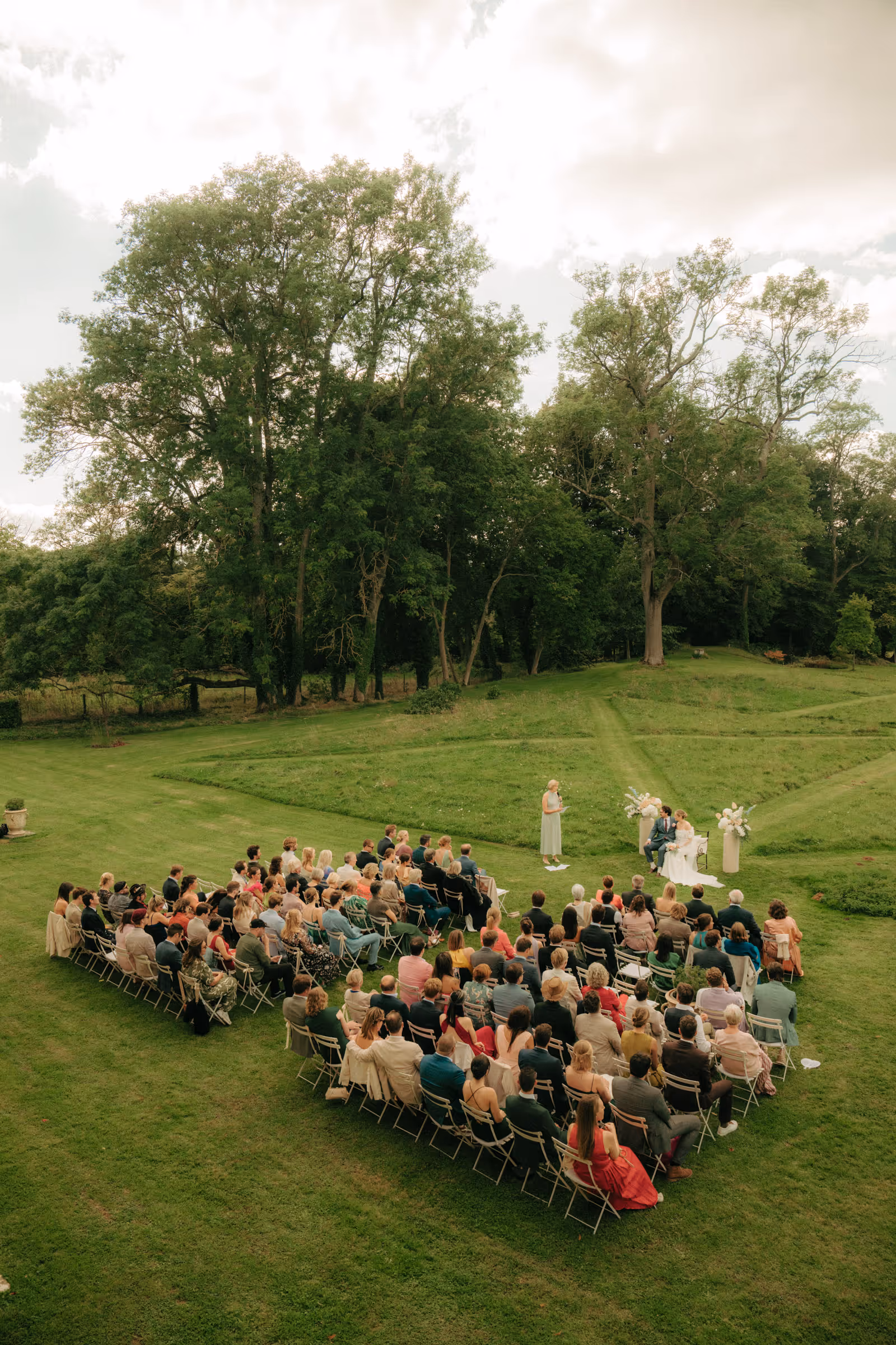 Outdoor wedding ceremony with guests seated around a bride and groom on a grassy lawn surrounded by trees.