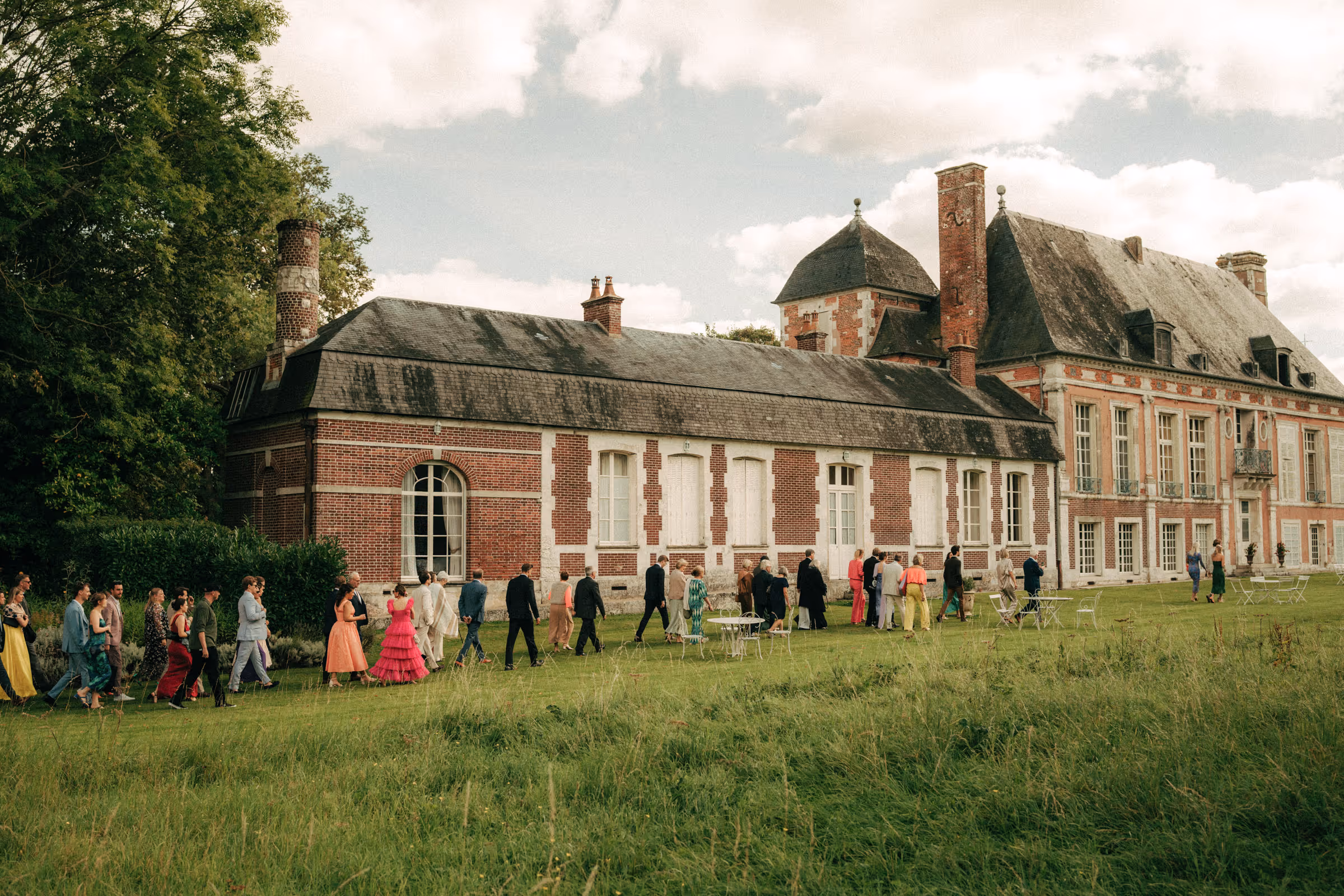 A group of people dressed in colorful formal attire walking on grass beside a large historic brick mansion with tall chimneys.