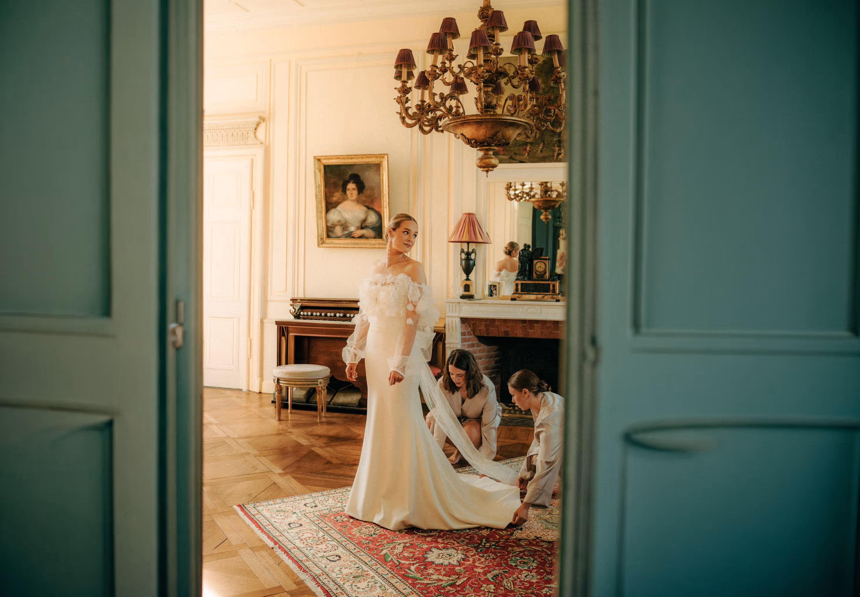 Bride standing in an elegant room with two women adjusting the train of her off-shoulder wedding dress.
