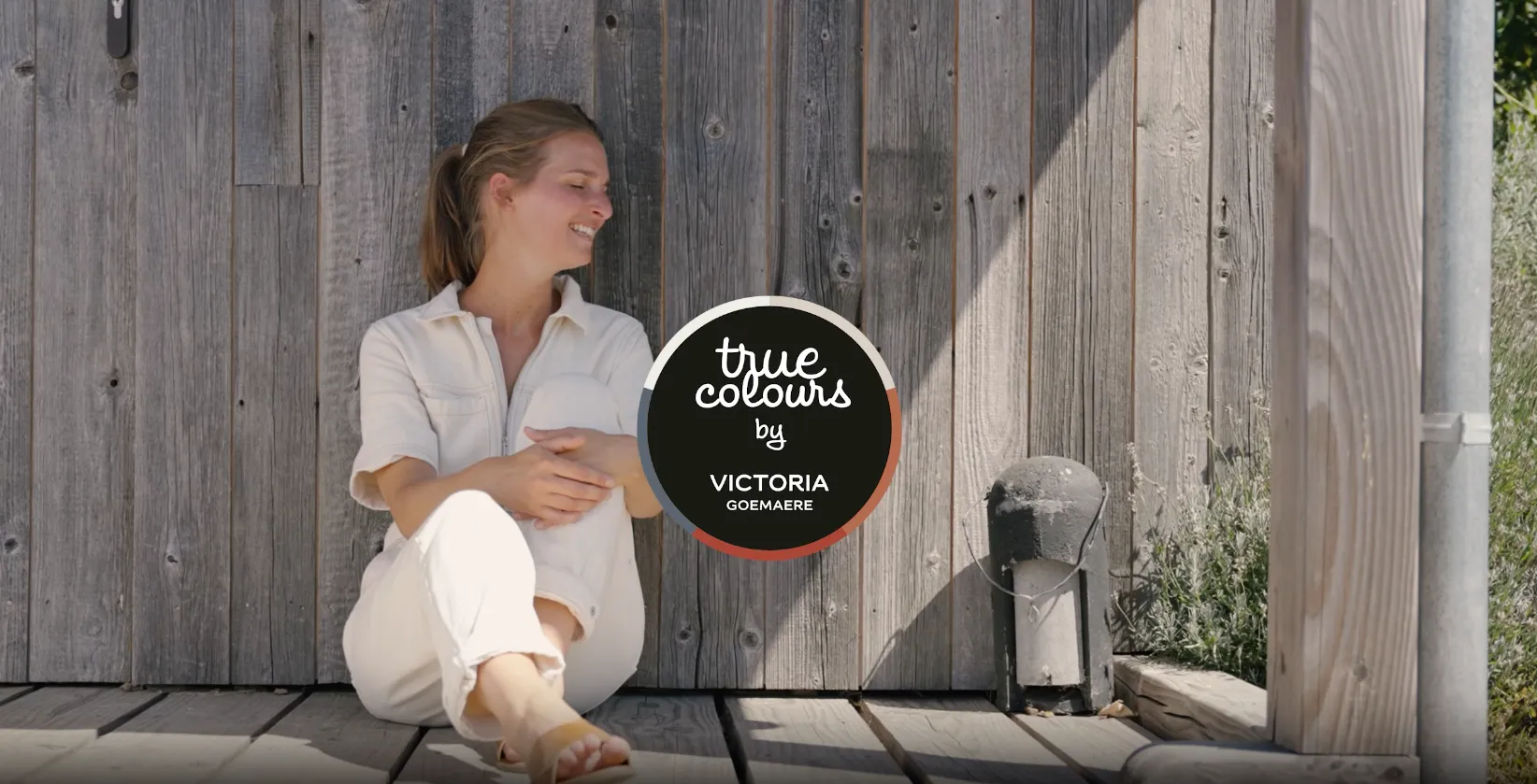 Woman in white sitting and smiling beside a rustic wooden wall with a circular logo for true colours by Victoria Goemaere.