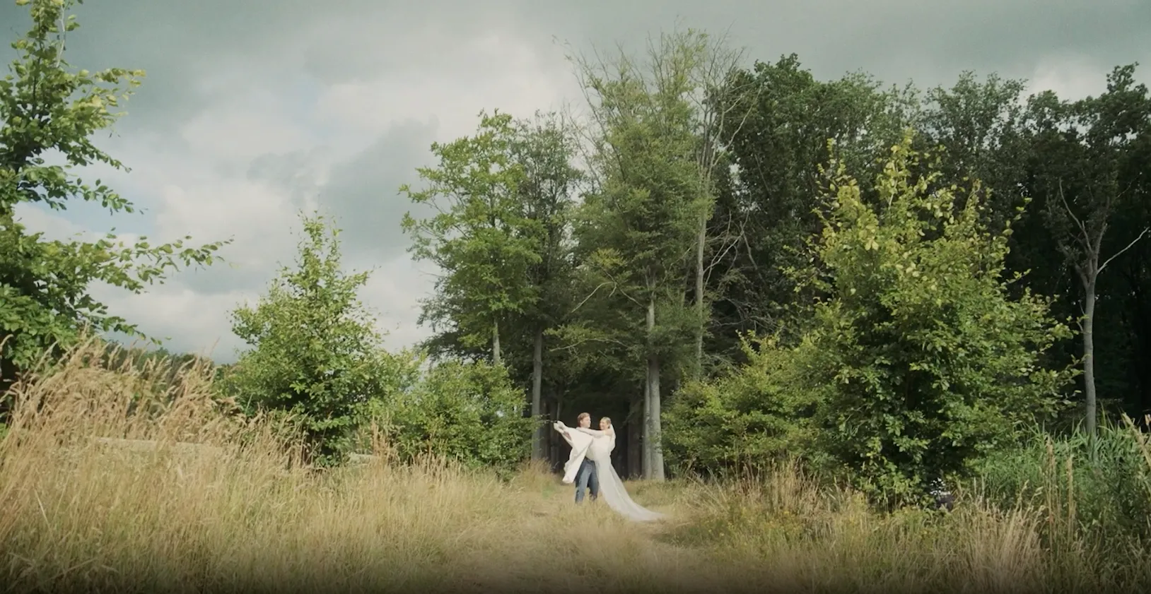 Couple in wedding attire dancing on a grassy path surrounded by tall trees and greenery under a cloudy sky.