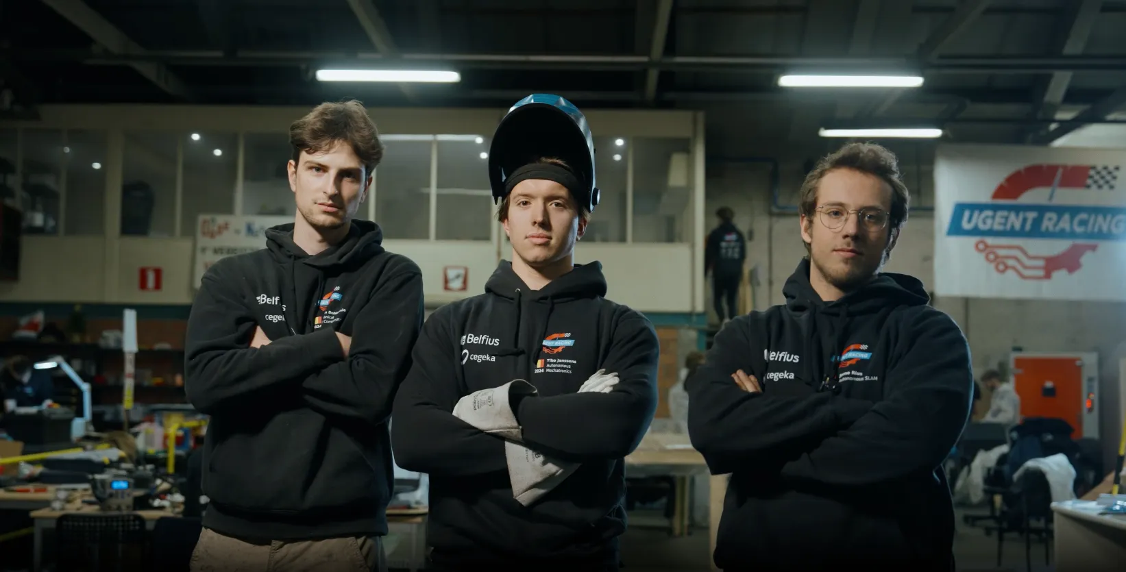 Three men in black hoodies posing with arms crossed in a workshop, one wearing a welding helmet lifted above his head.