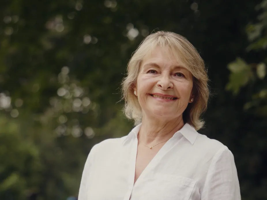 Smiling older woman with short blond hair wearing a white shirt against a blurred green foliage background.