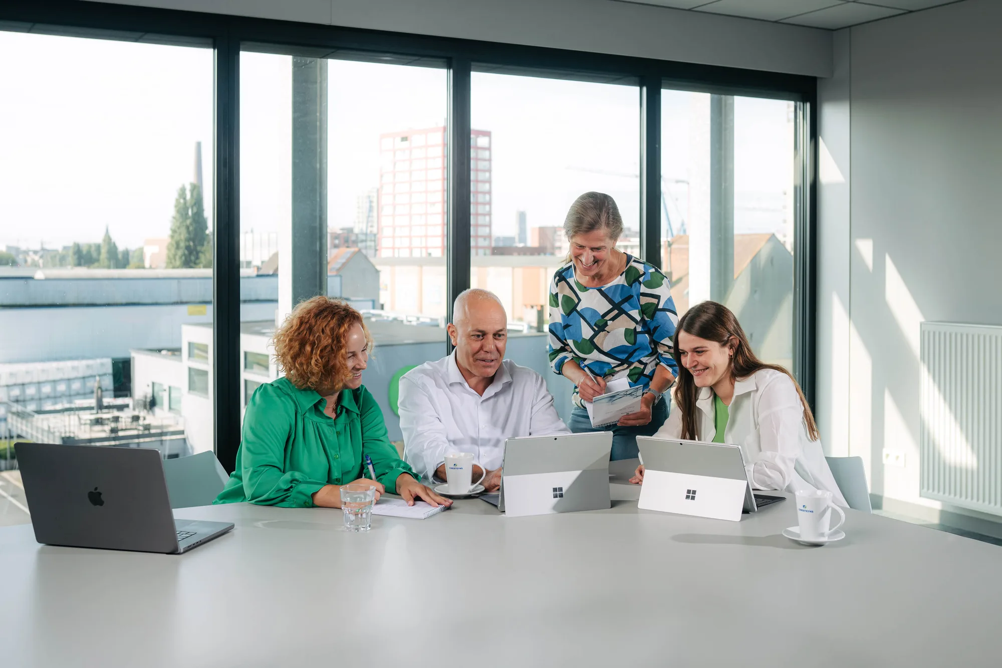 Four colleagues collaborating around a table in a bright office with laptops, coffee mugs, and a notepad.