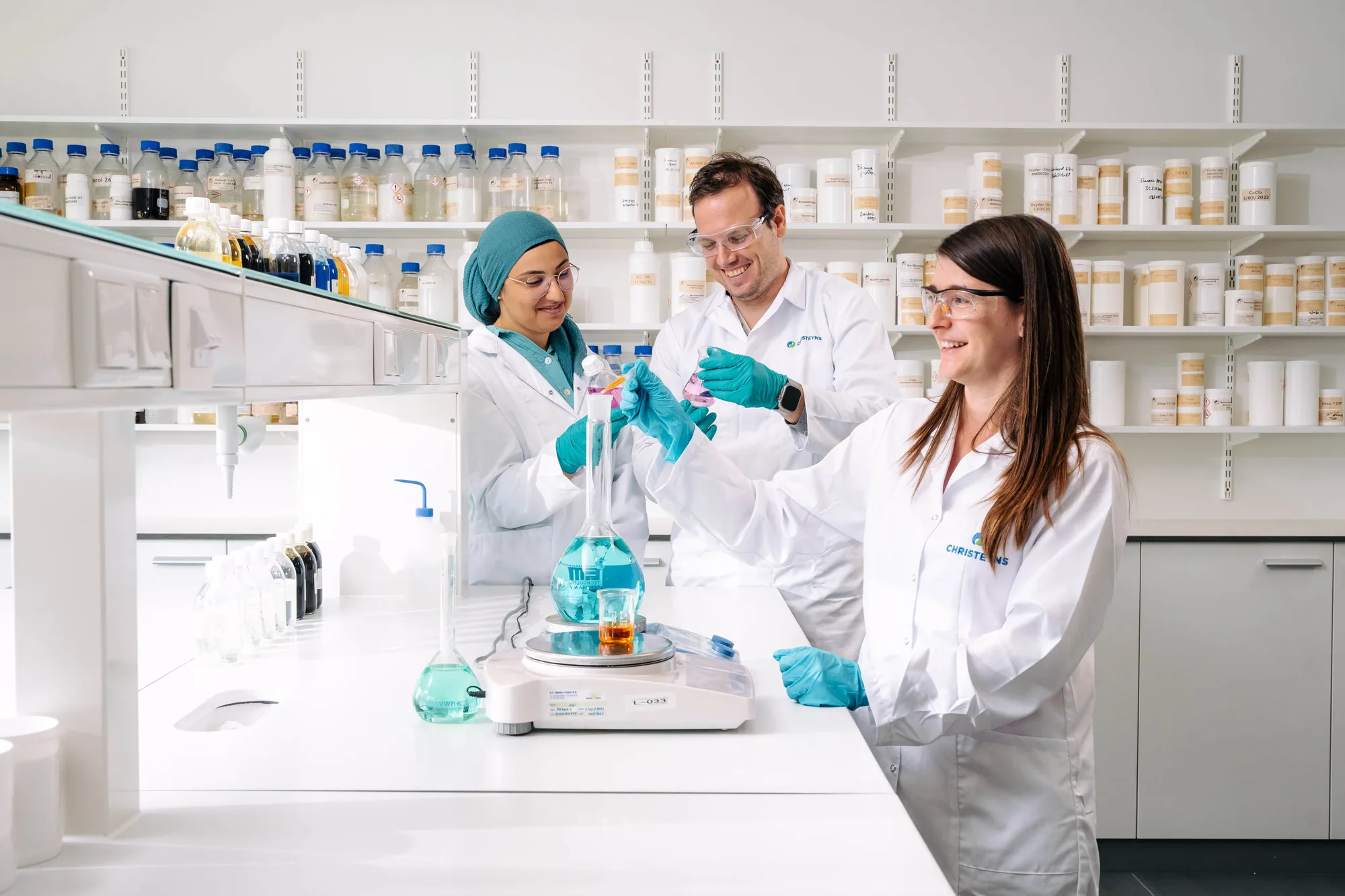 Three scientists wearing white lab coats and safety glasses working with blue and orange liquids in a bright laboratory.