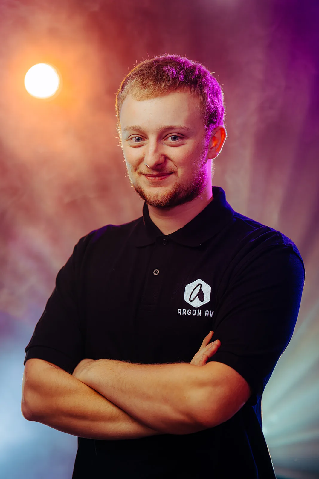 Smiling man with short blond hair wearing a black Argon AV polo shirt, standing with arms crossed against a colorful background with orange and purple lighting.