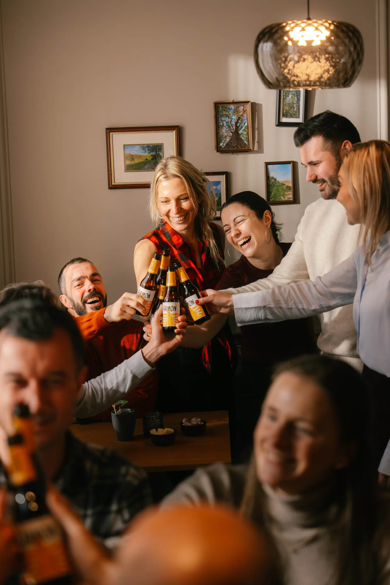 Group of friends smiling and clinking beer bottles together indoors under a hanging light.