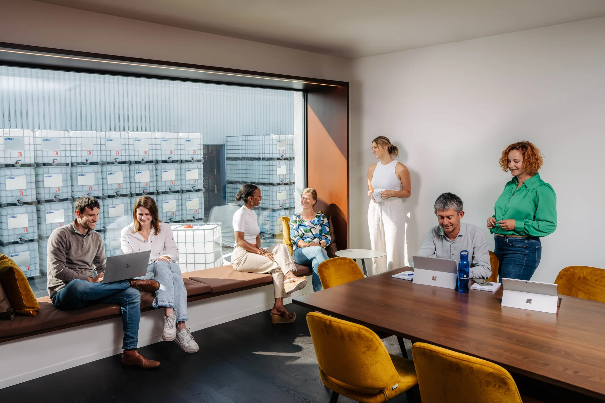 Six diverse colleagues casually interacting and working with laptops and tablets in a bright office with large window and mustard yellow chairs.
