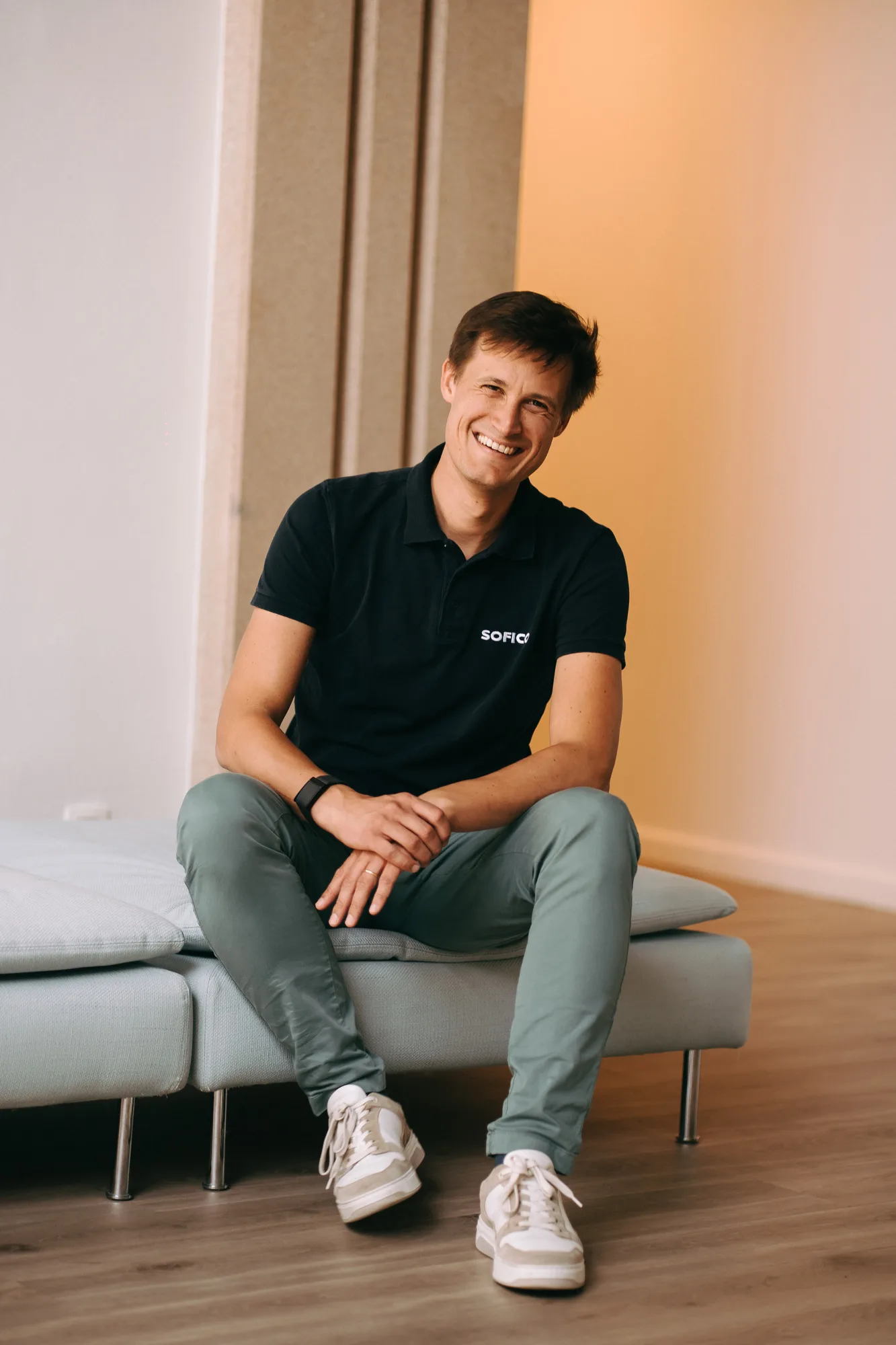Smiling man in a black polo shirt with 'SOFICO' logo, green pants, and white sneakers sitting on a modern light gray sofa.