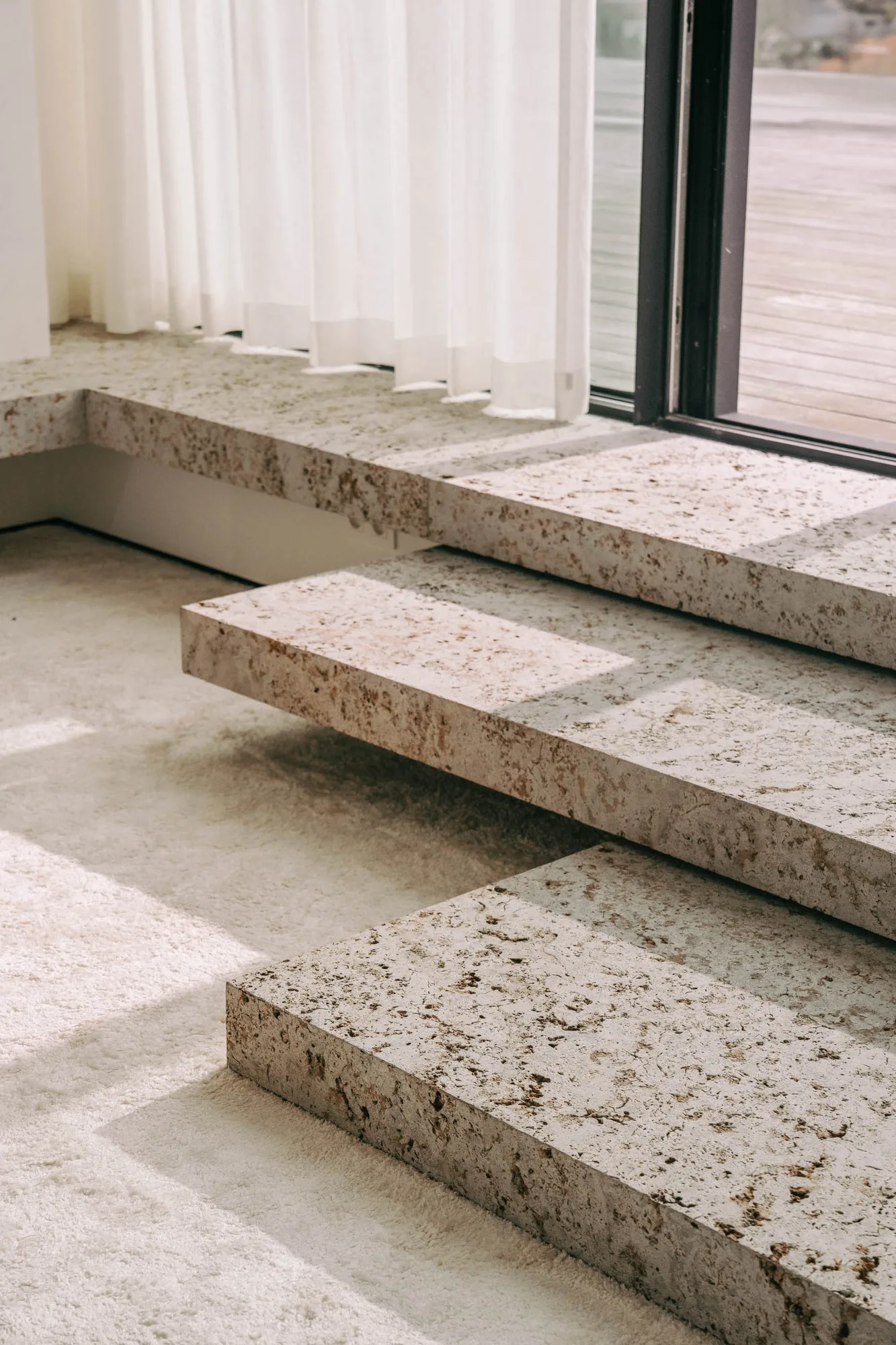 Modern interior showing textured stone steps next to a large window with sheer white curtains and sunlight casting shadows.