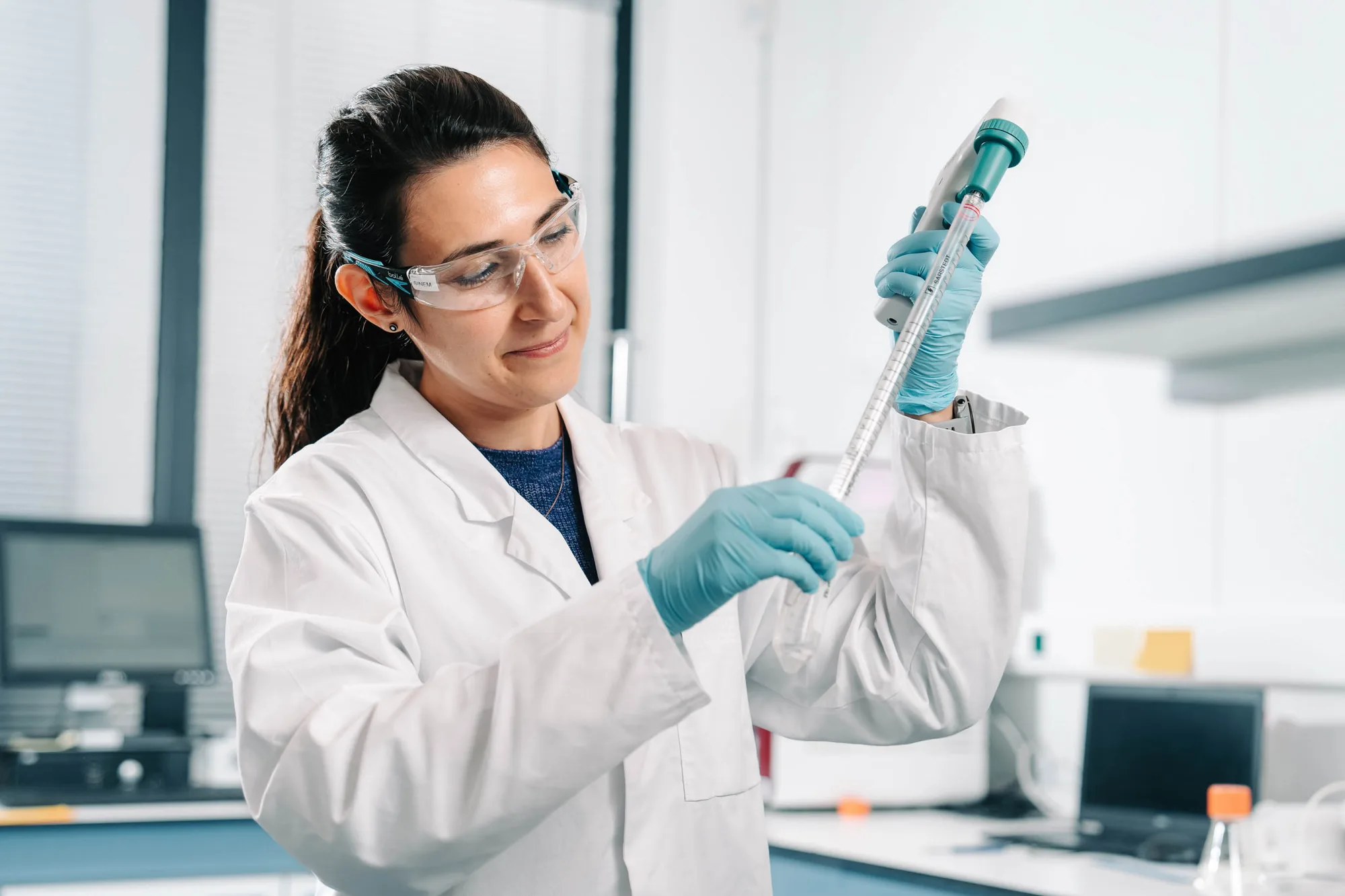 Female scientist in a lab coat and safety glasses using a pipette to measure liquid in a laboratory.