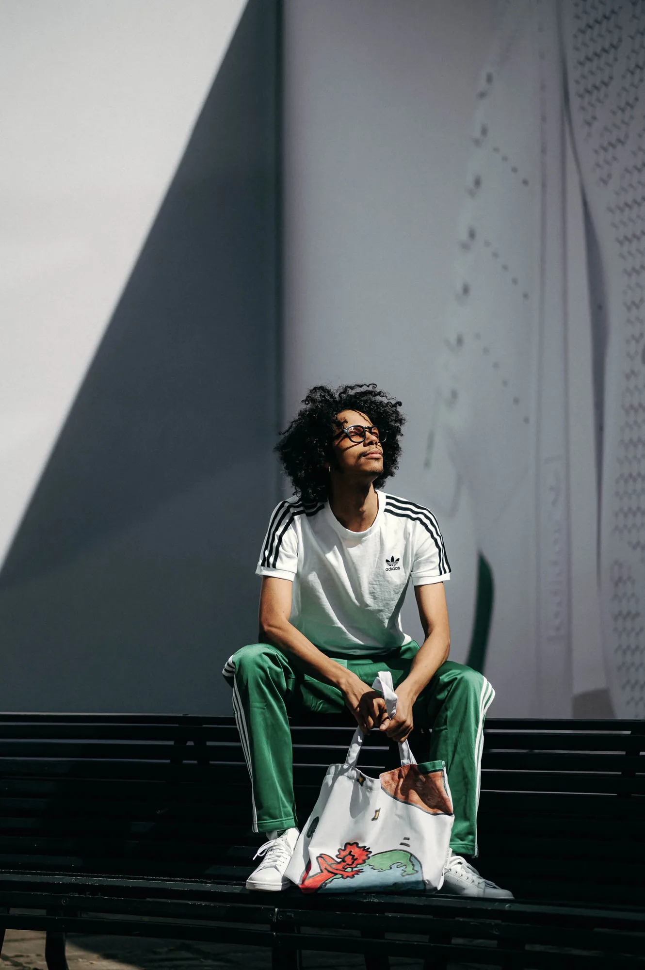Man in green Adidas track pants and white Adidas t-shirt sitting on a bench holding a colorful tote bag.