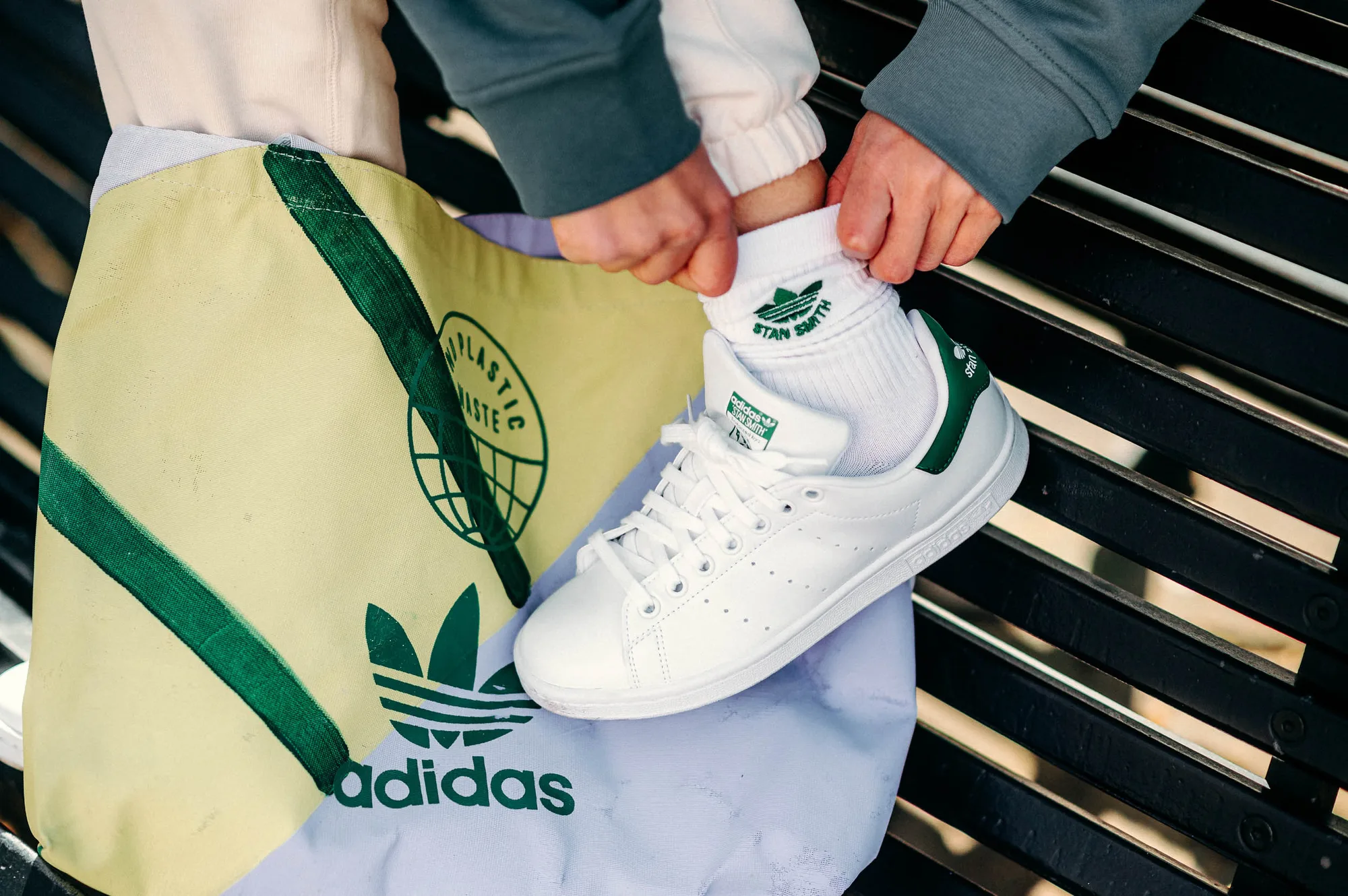 Person wearing white Adidas Stan Smith sneakers and white socks with Stan Smith logo, adjusting the sock while sitting on a black bench next to a light yellow and light purple Adidas tote bag.