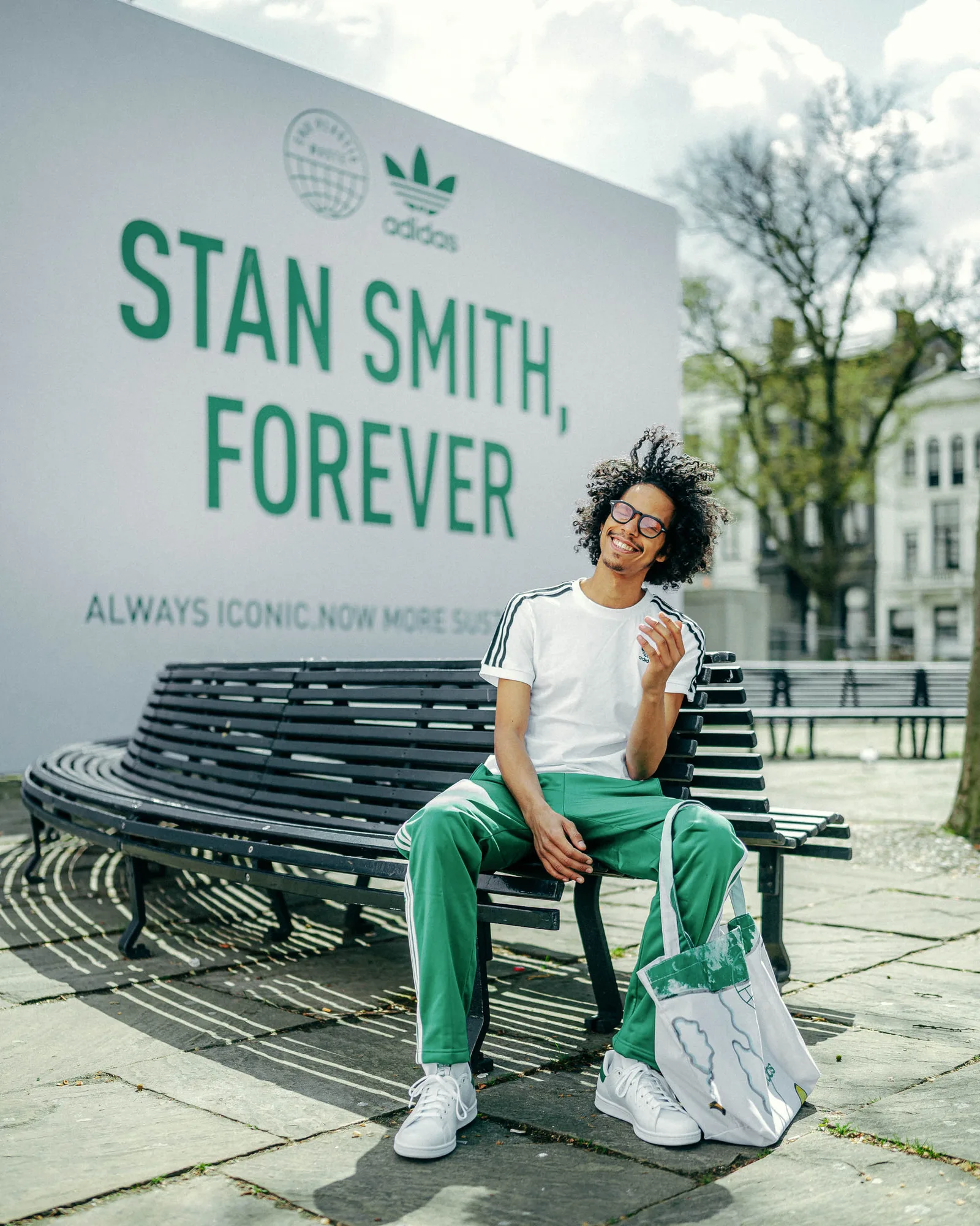 Smiling man with curly hair wearing Adidas white t-shirt and green pants sitting on a curved bench outdoors with a tote bag beside him and a large 'Stan Smith, Forever' Adidas advertisement in the background.