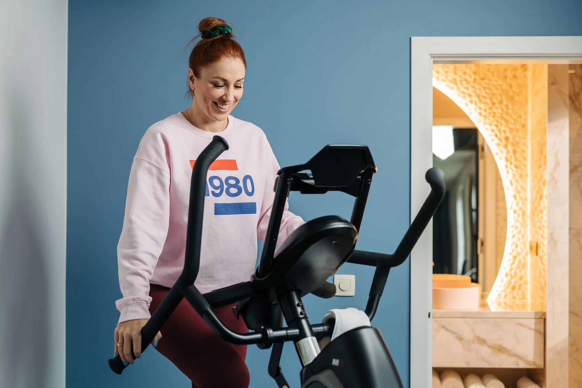 Smiling woman in pink sweatshirt using a stationary exercise bike indoors with blue wall and a lit mirror in the background.