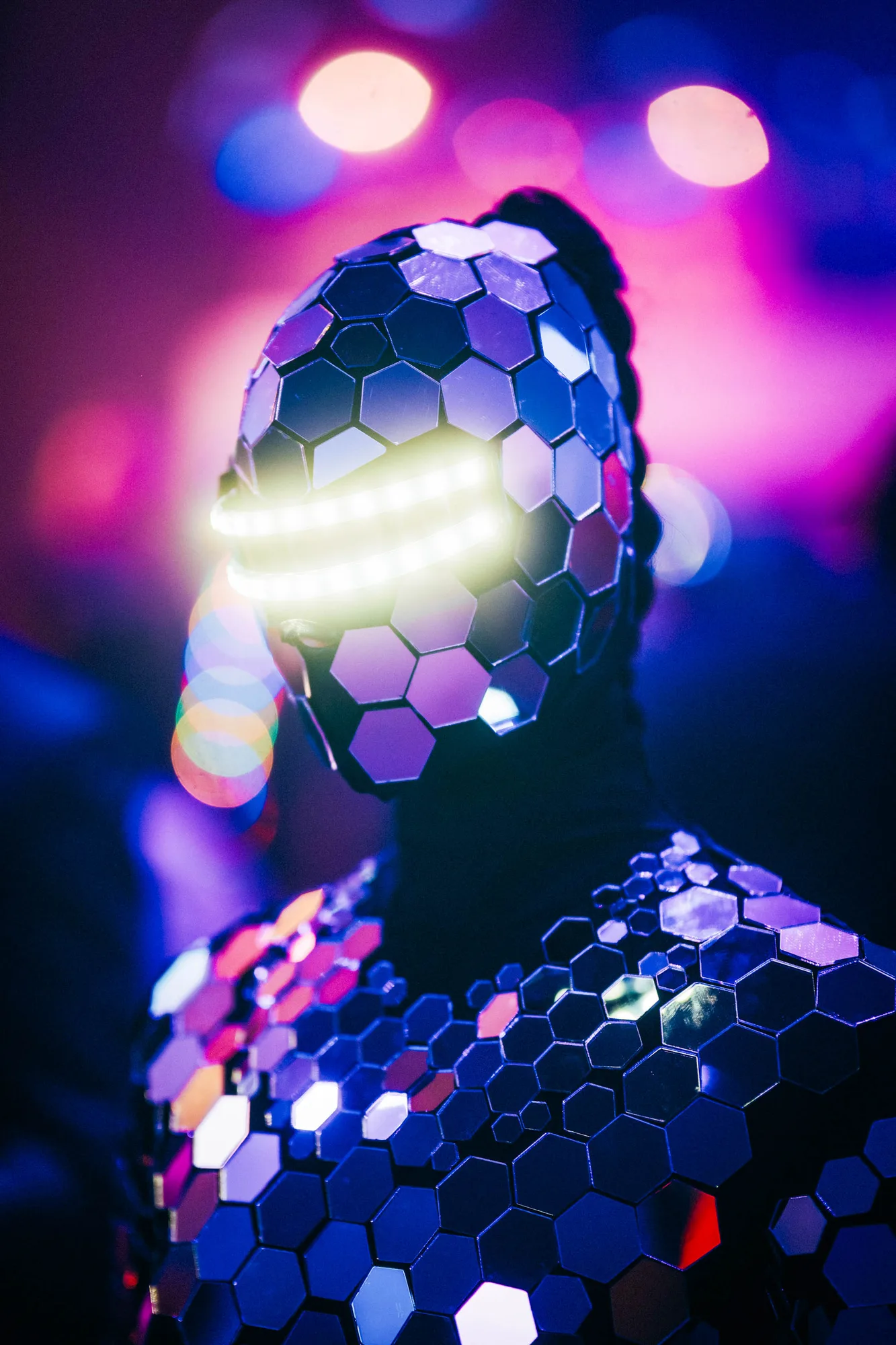 Person wearing a futuristic costume covered in reflective hexagonal mirrors with glowing horizontal light bars over the eyes against a purple and blue blurred background.