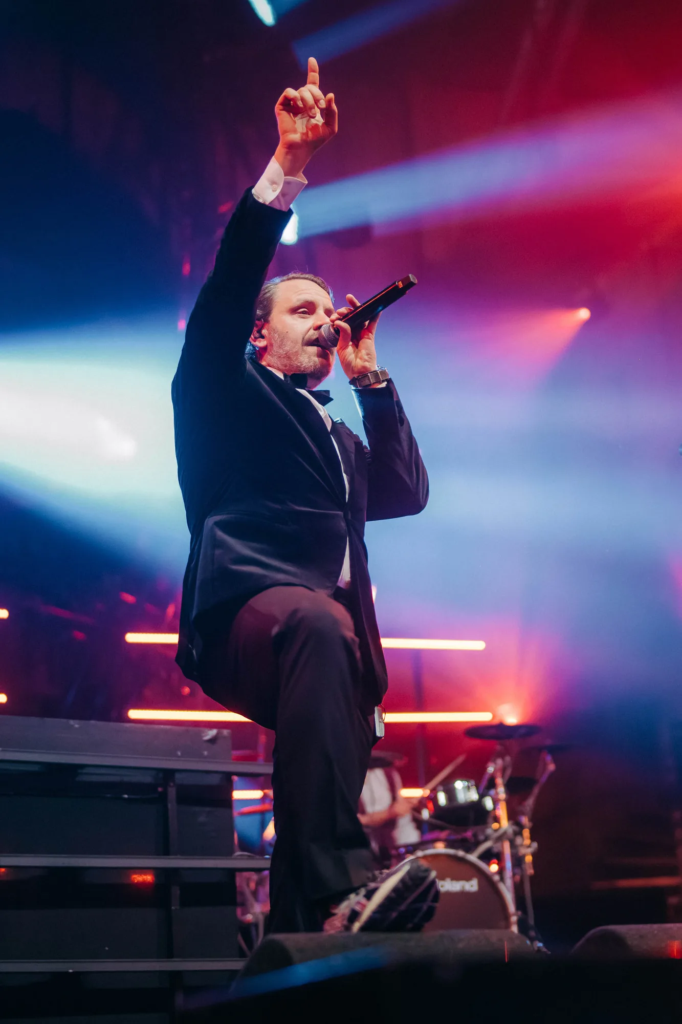 Male singer in black suit performing on stage with microphone, colorful stage lights and drummer in the background.