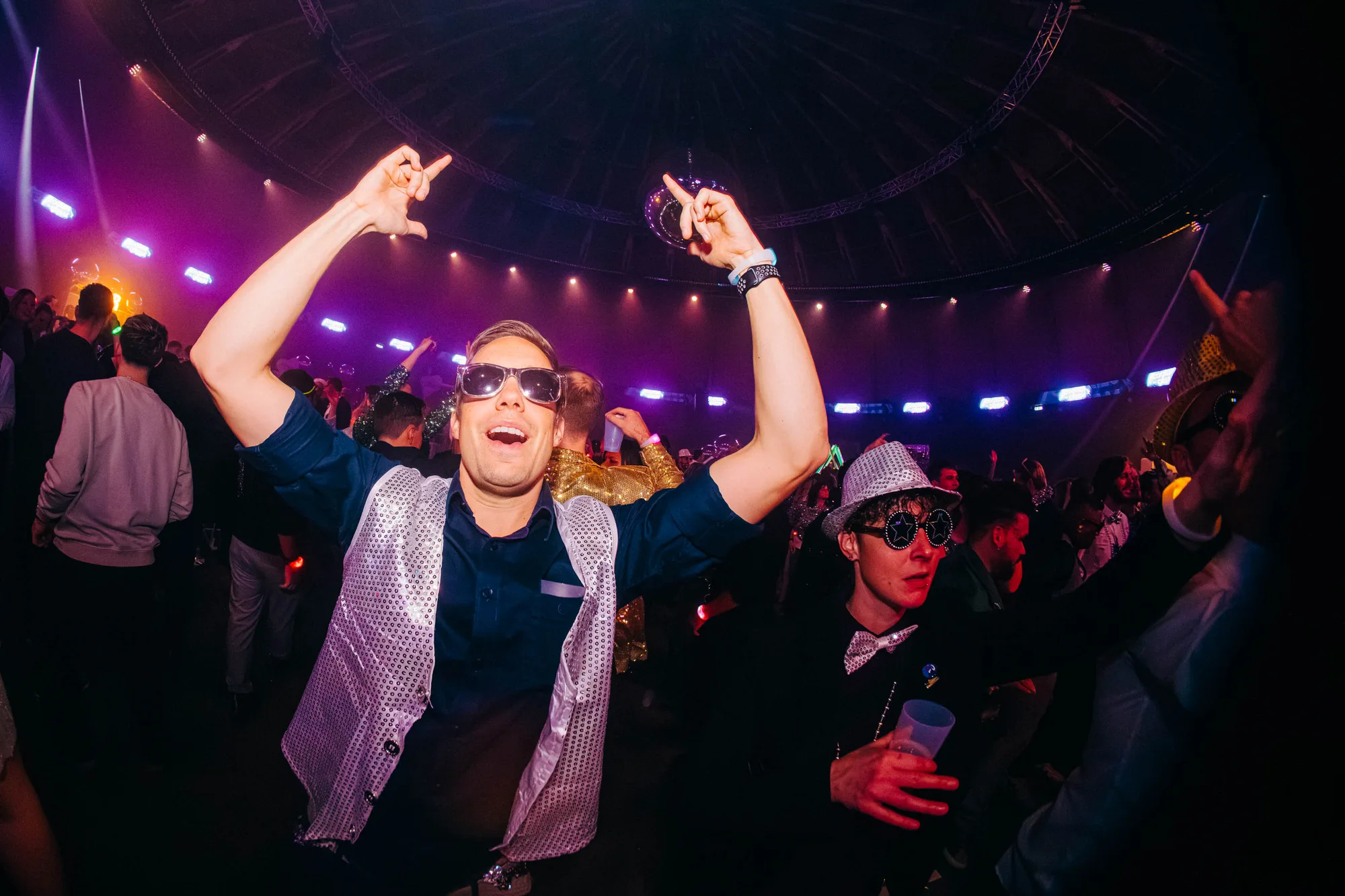 Two men in festive attire dancing at a crowded indoor party with colorful lights and a disco ball overhead.