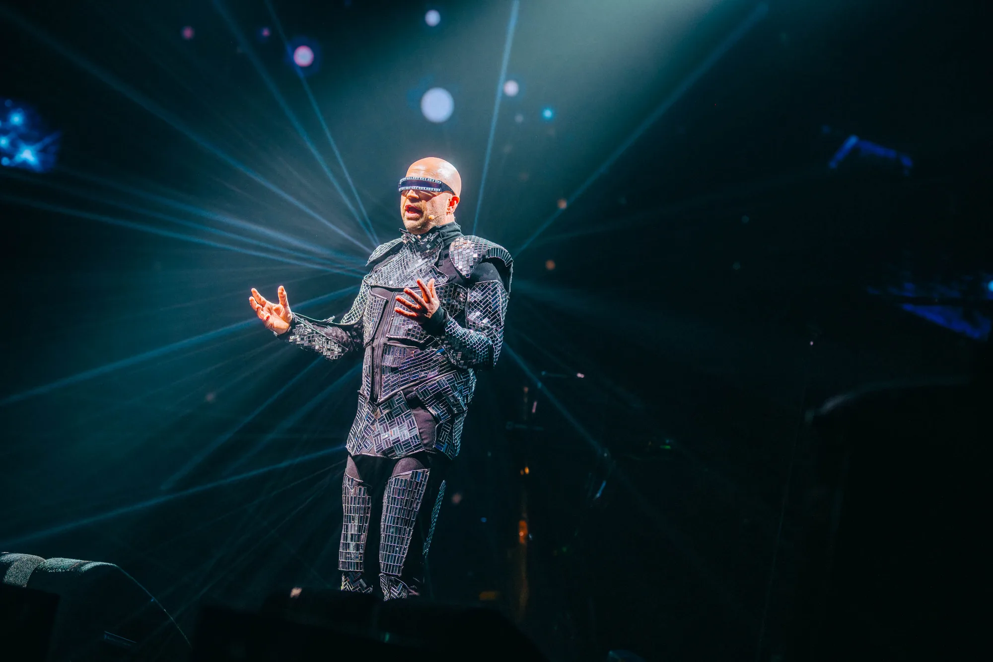 Performer in futuristic costume and visor singing on stage with blue light beams radiating behind.