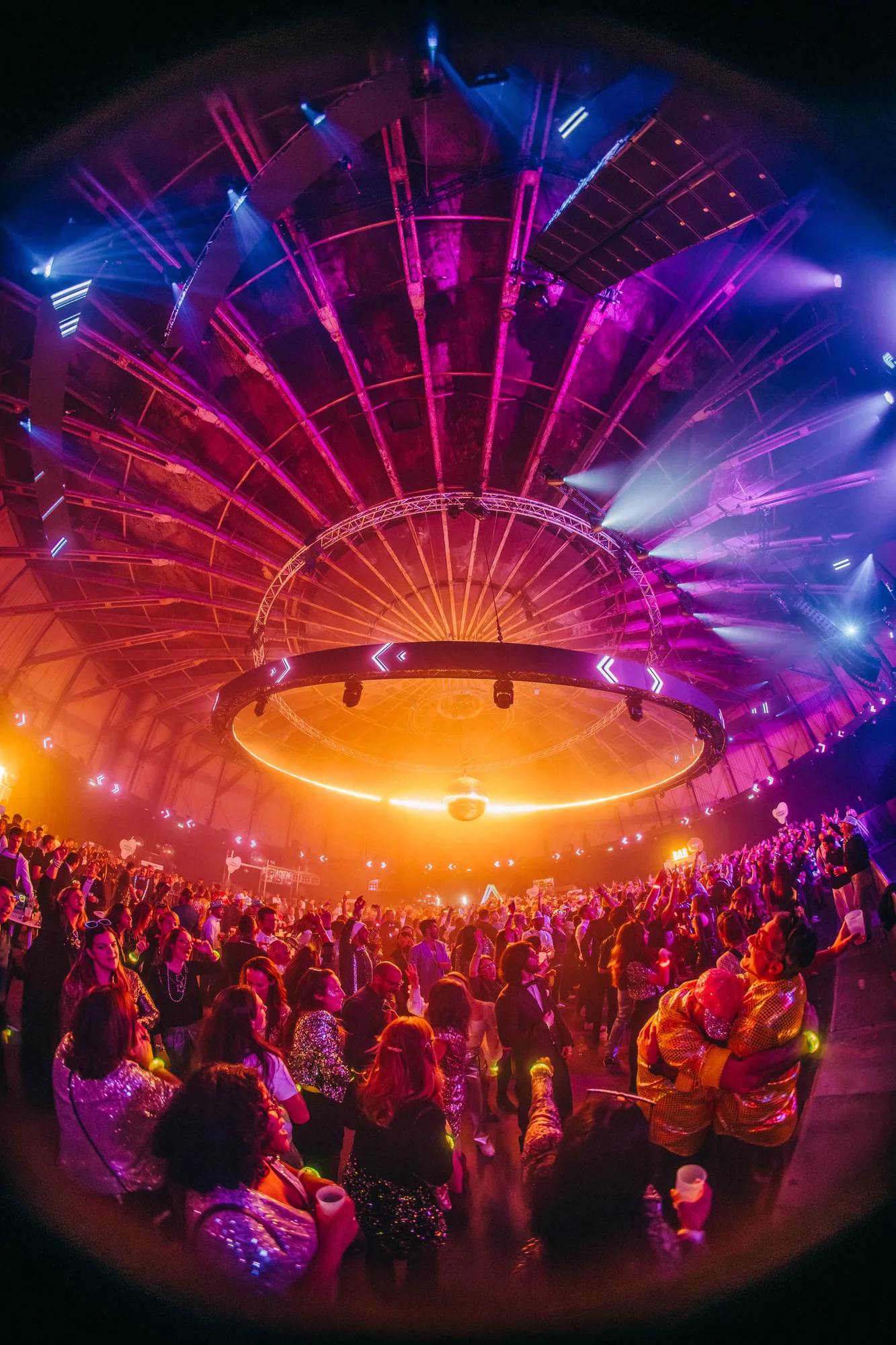Large crowd dancing and socializing under a glowing circular light fixture inside a dome-shaped venue with colorful stage lighting.