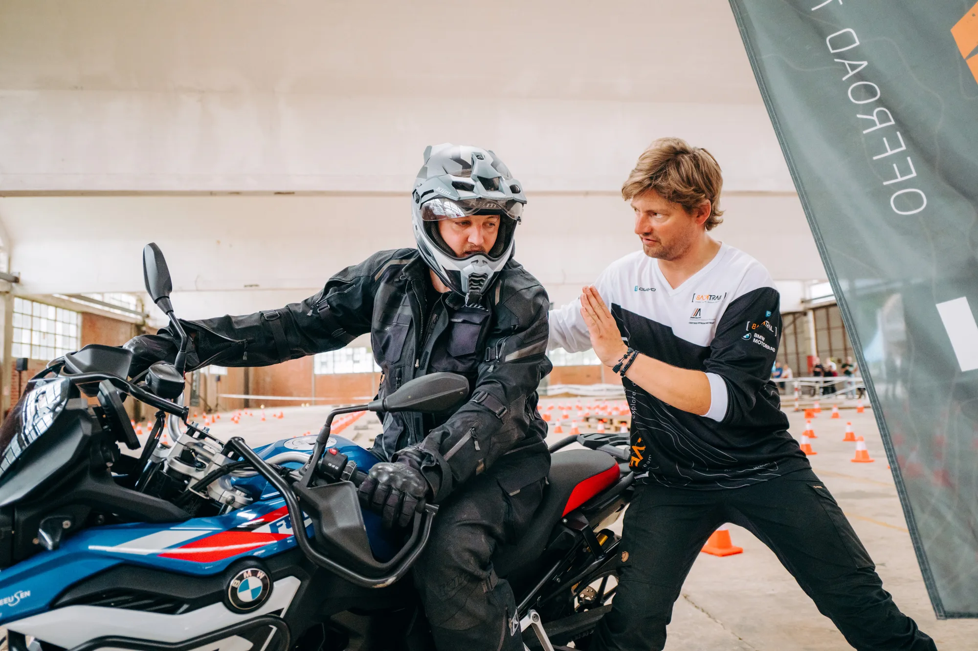 Motorcycle instructor guiding a rider dressed in protective gear on a blue BMW motorcycle inside a training facility.