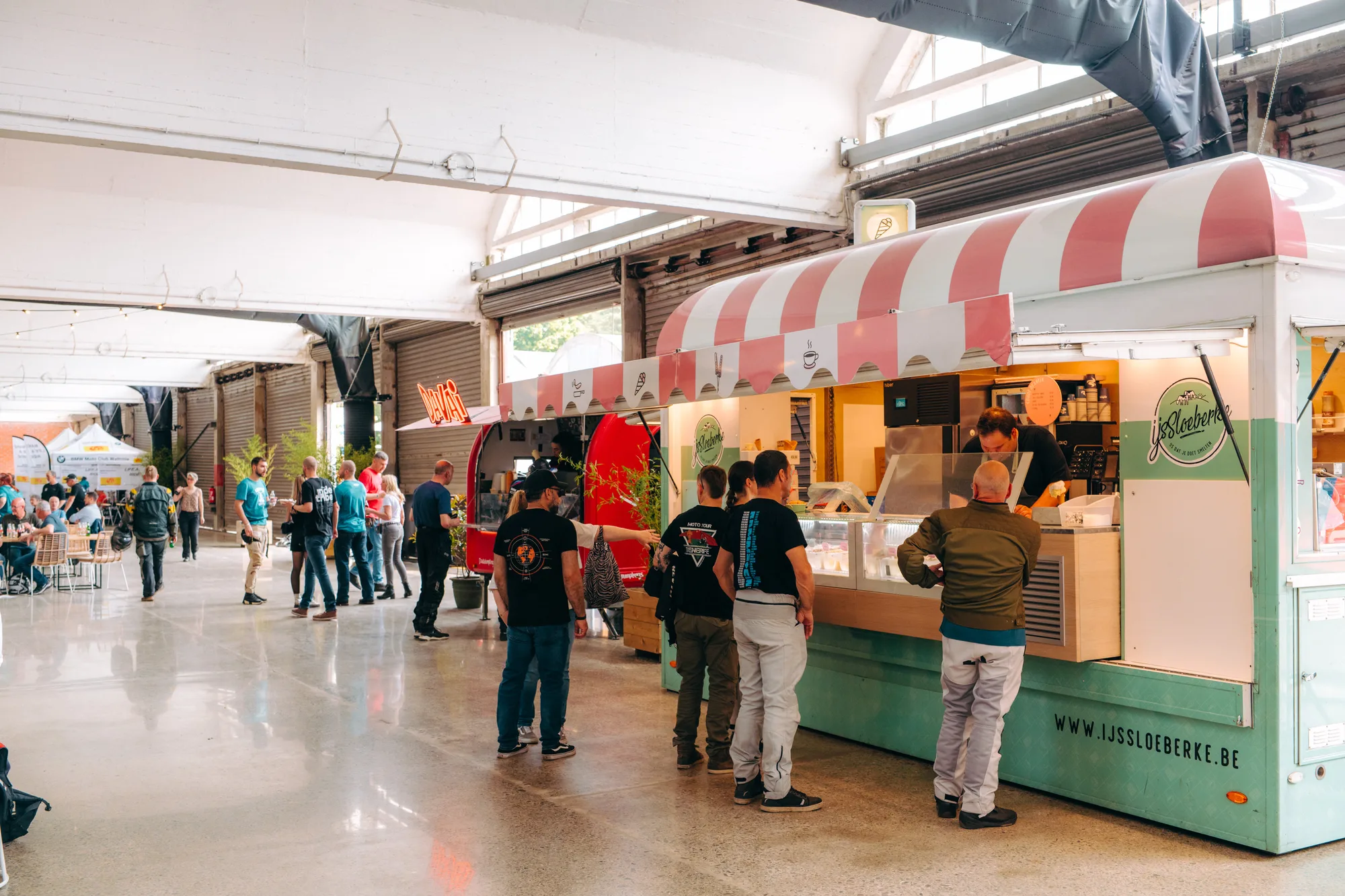 People standing in line at a pastel green and pink striped ice cream food truck inside a large indoor market.