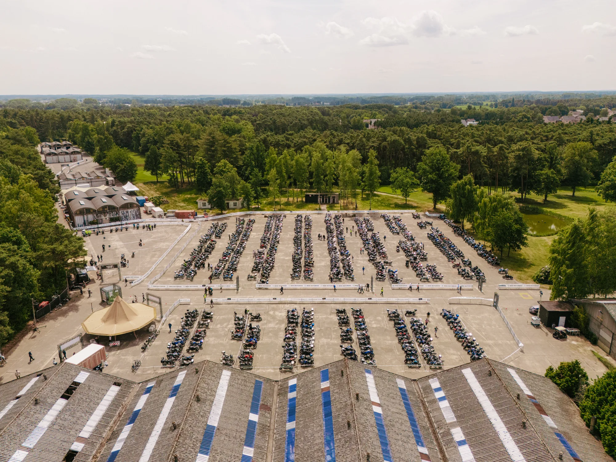 Aerial view of a large outdoor parking area filled with motorcycles arranged in rows, surrounded by trees and buildings.