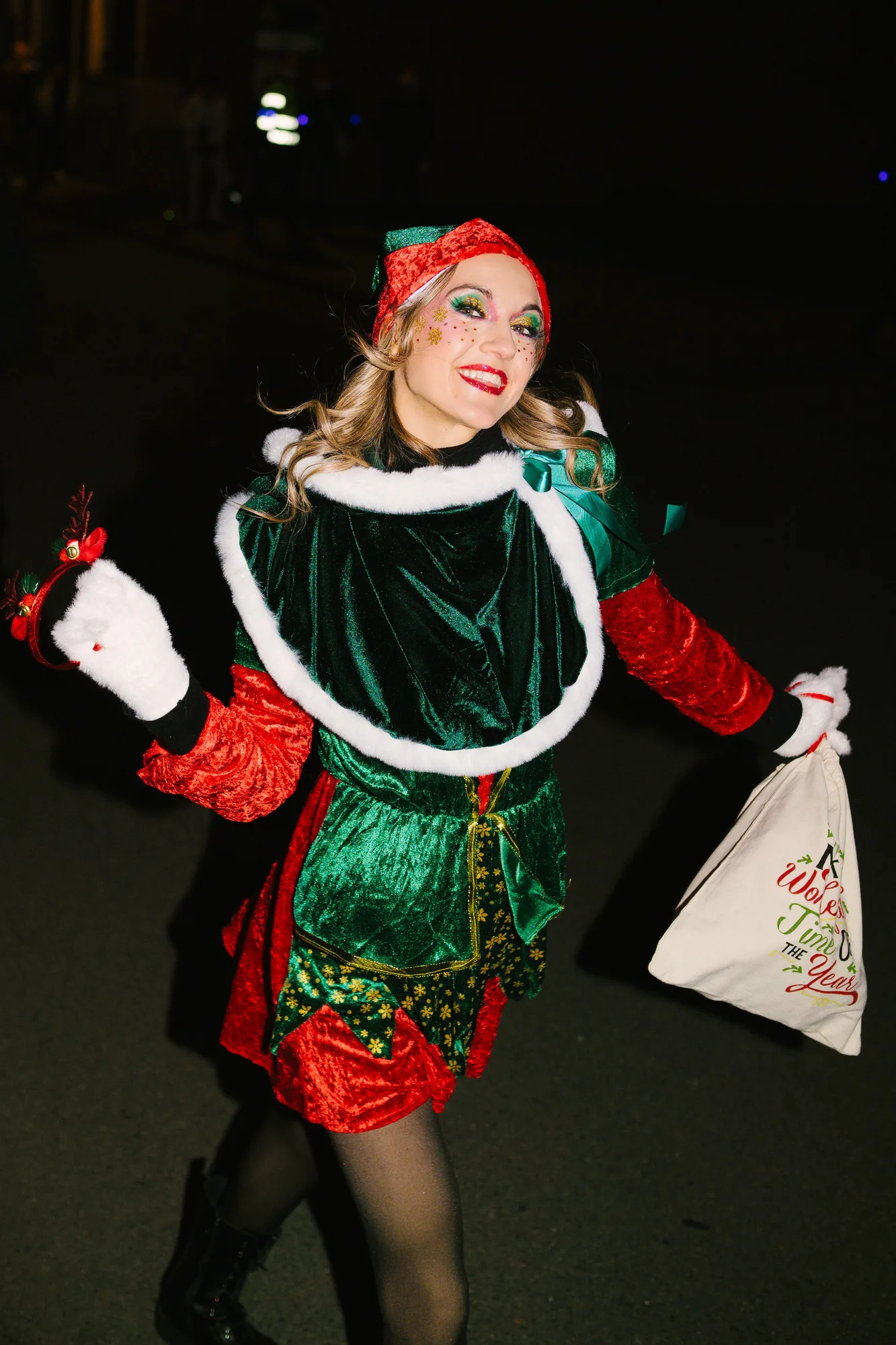 Smiling woman dressed in a festive elf costume with green and red velvet, holding a holiday-themed bag and reindeer antler headband.