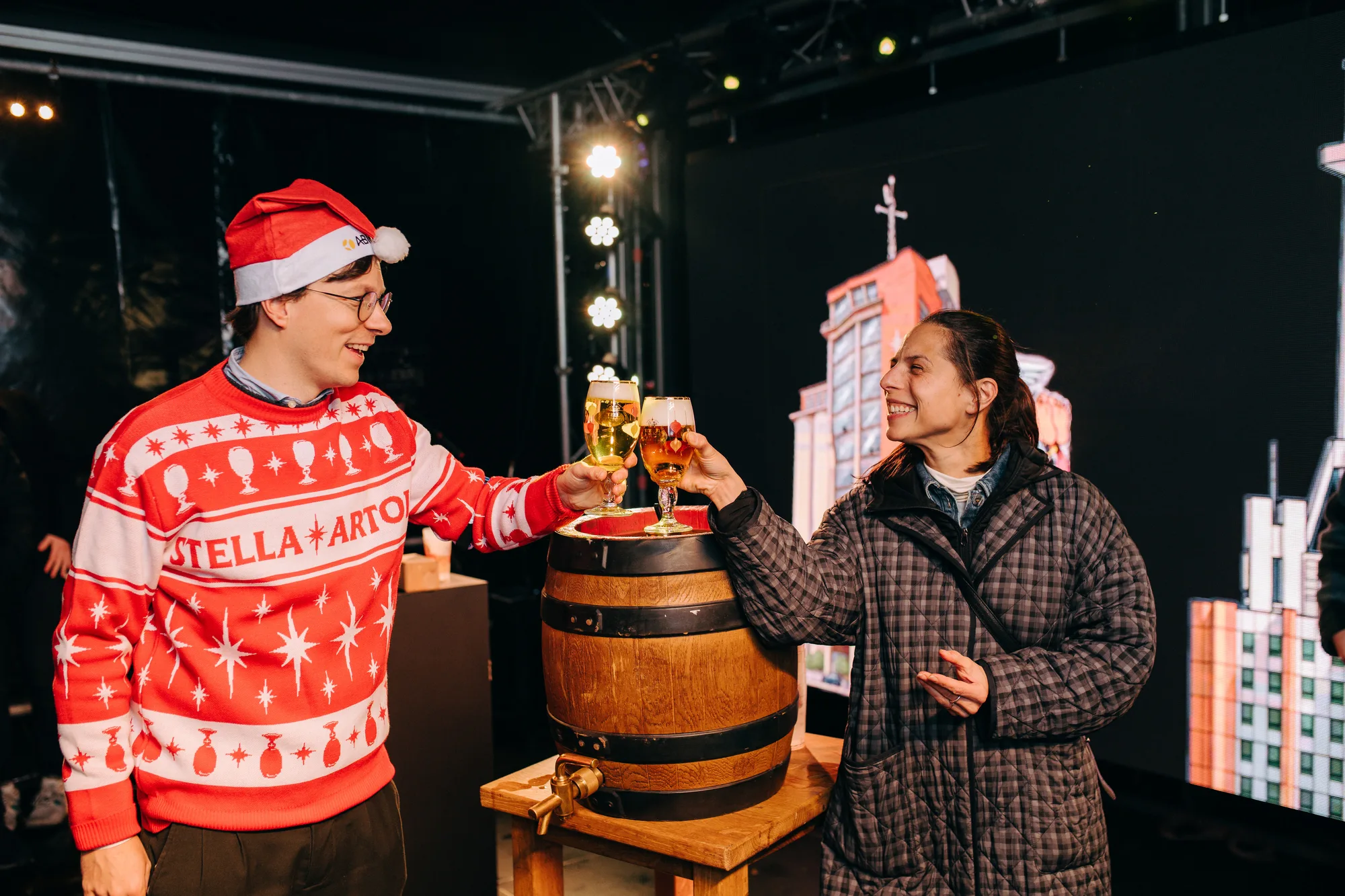 Two people smiling and clinking beer glasses next to a wooden beer barrel at an indoor event.