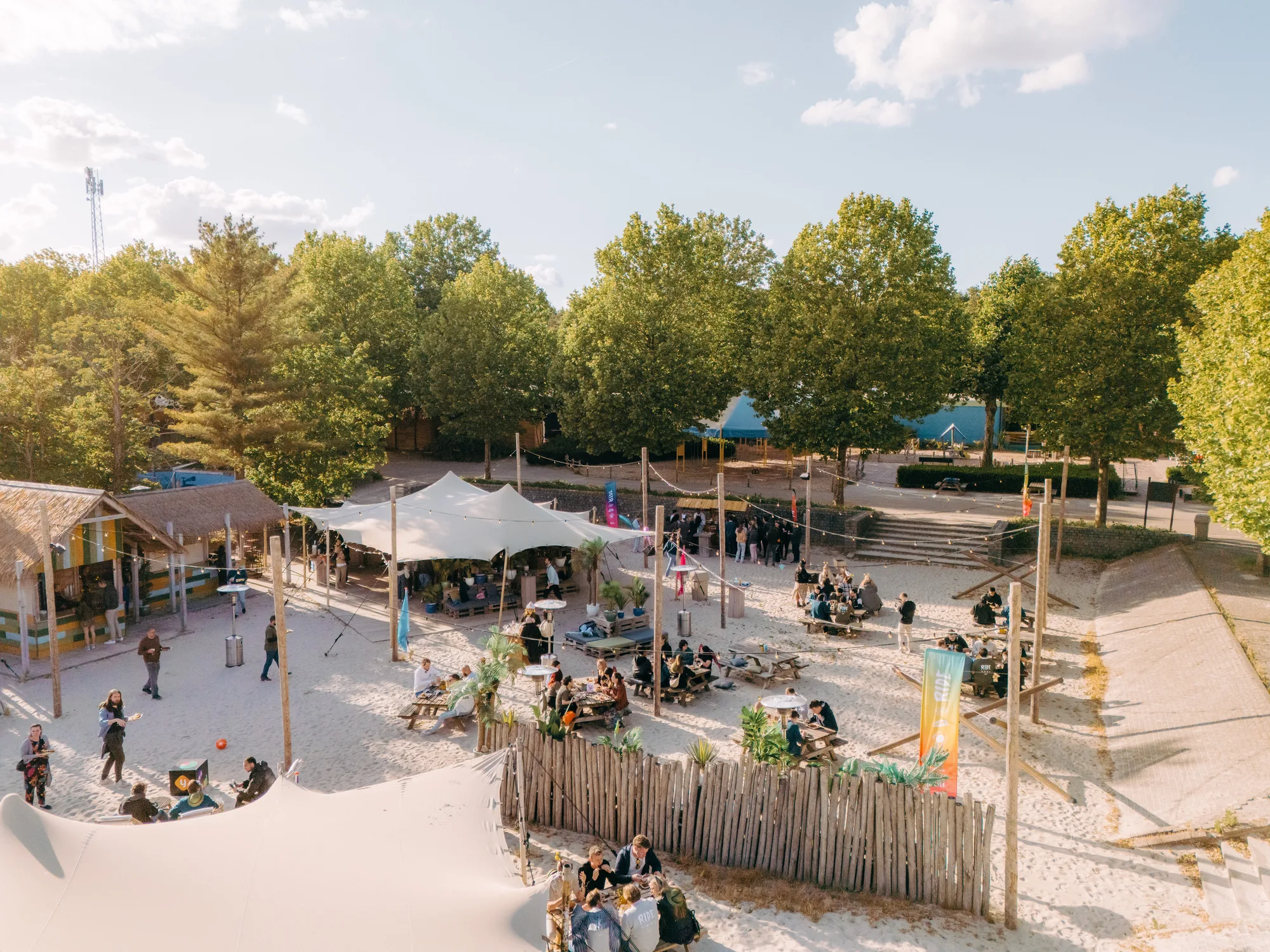 Outdoor beach-style event space with people sitting at picnic tables, tents, string lights, and surrounded by trees under a clear sky.