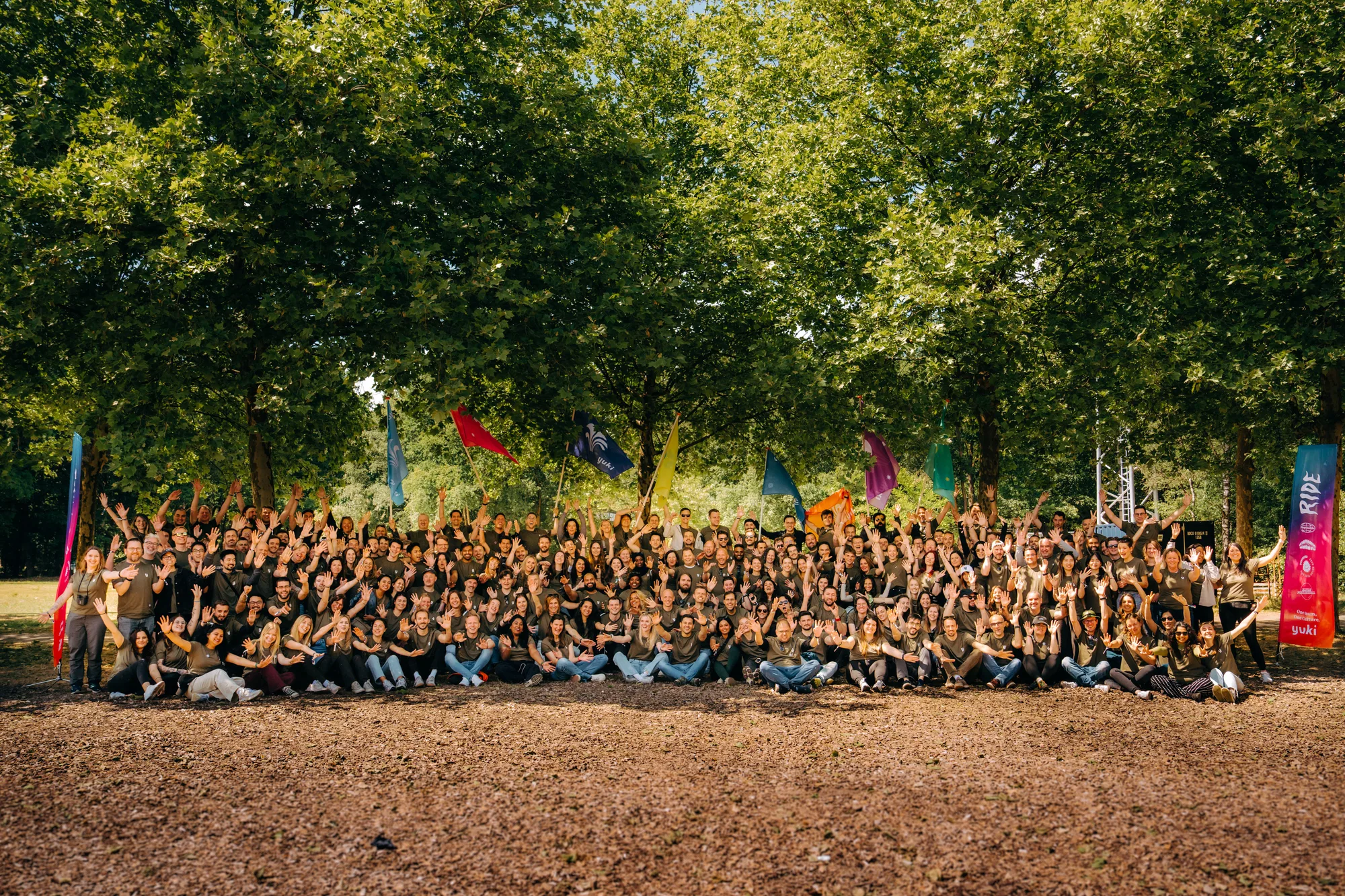Large group of people sitting and standing outdoors under trees, waving hands and holding colorful flags.