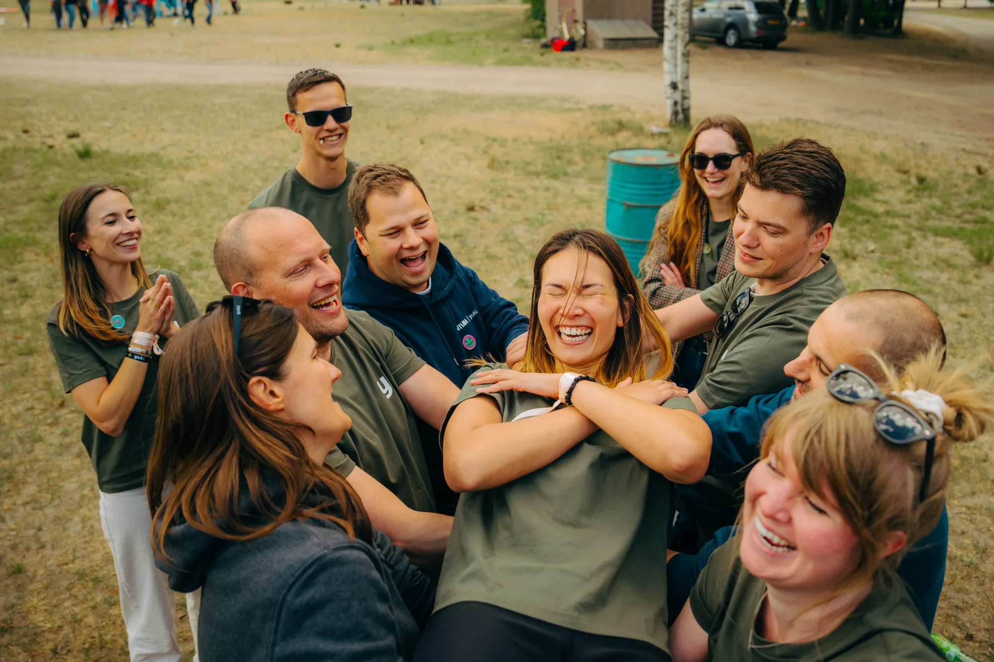 Group of smiling friends outdoors lifting a laughing woman in an arm-crossed pose during a team-building activity.