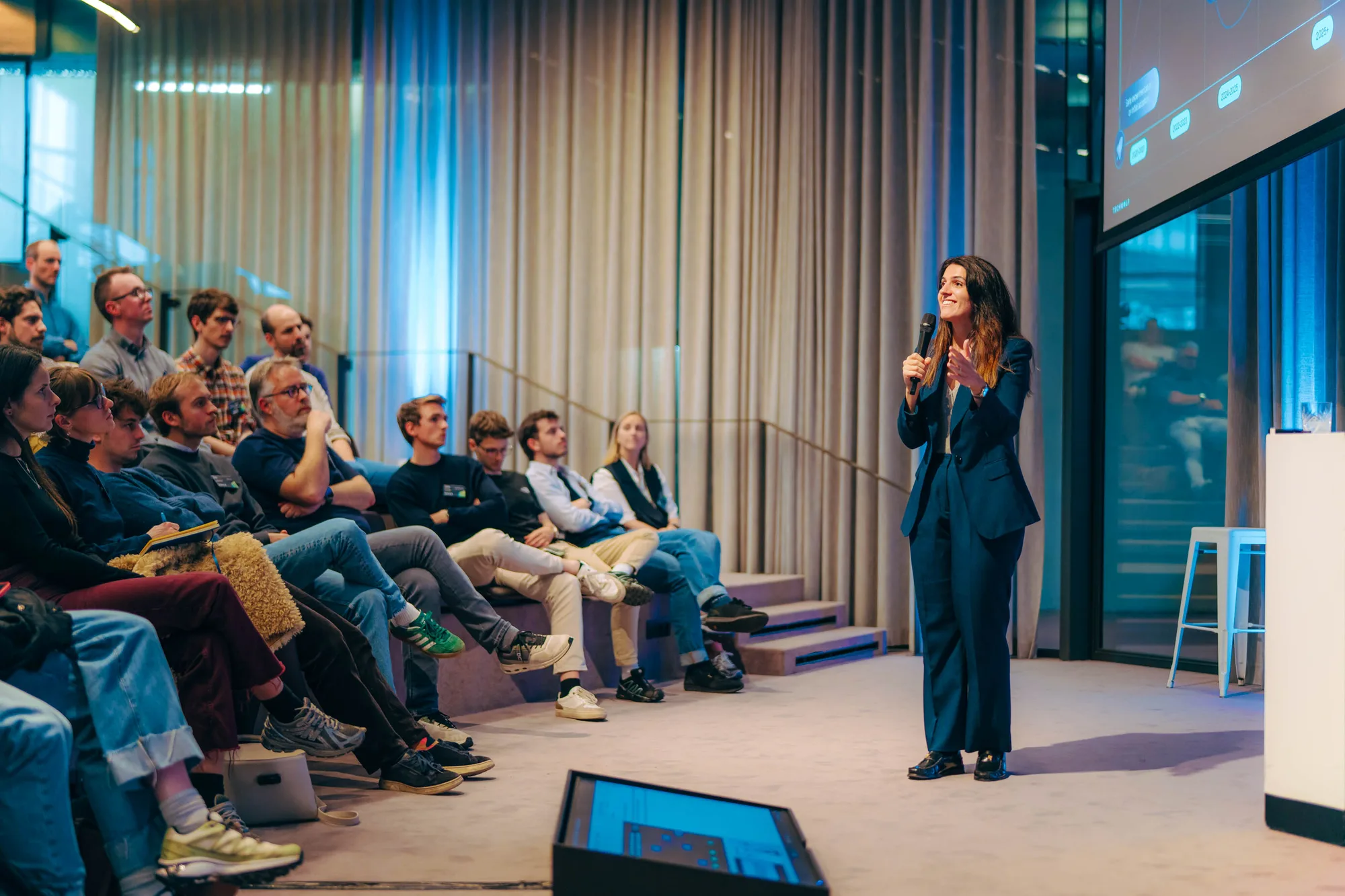 Female speaker in navy suit presenting with microphone to seated audience in modern conference room.