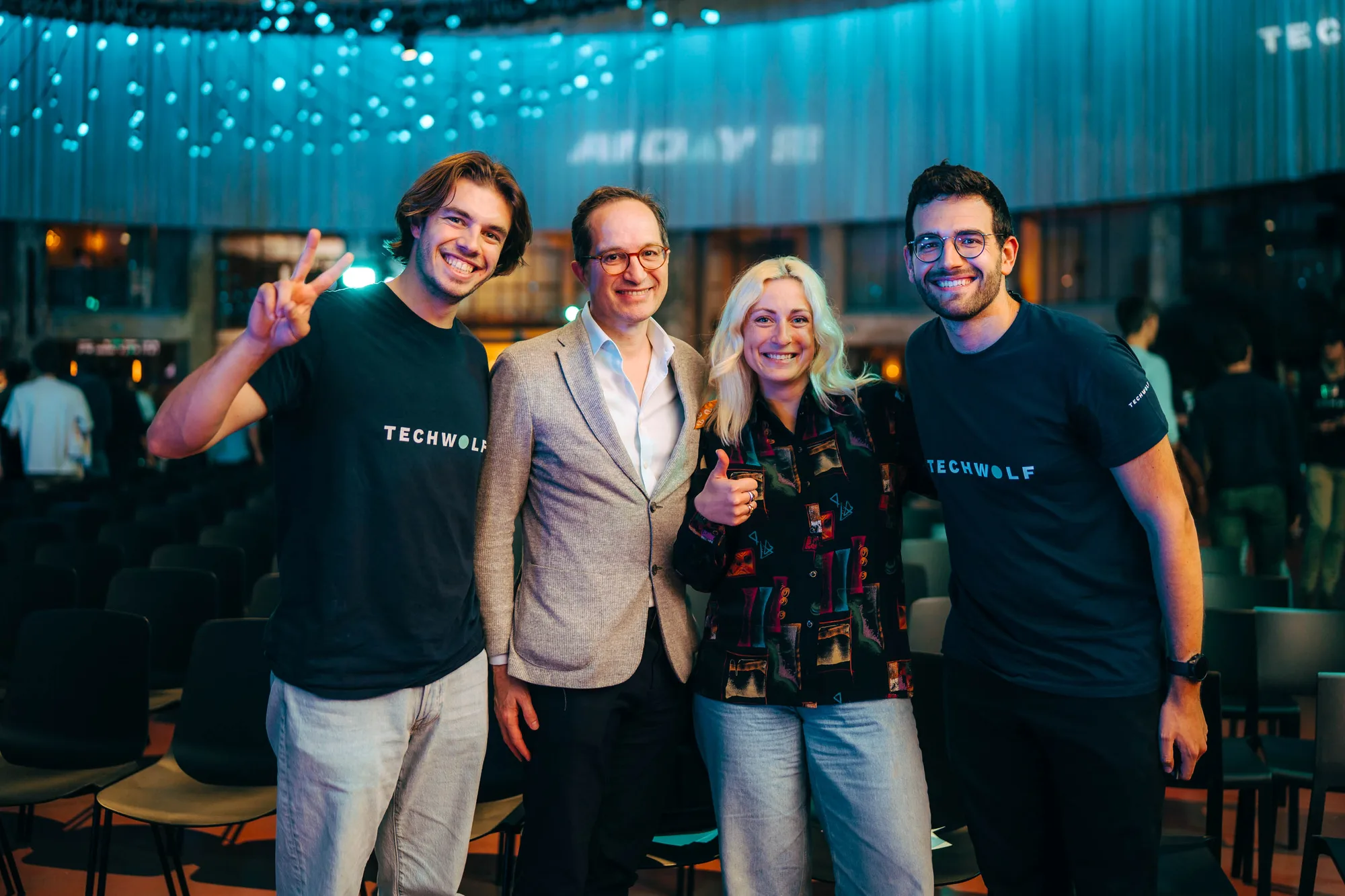 Four people smiling indoors with two men wearing TECHWOLF shirts, one man in a blazer, and one woman giving a thumbs up.
