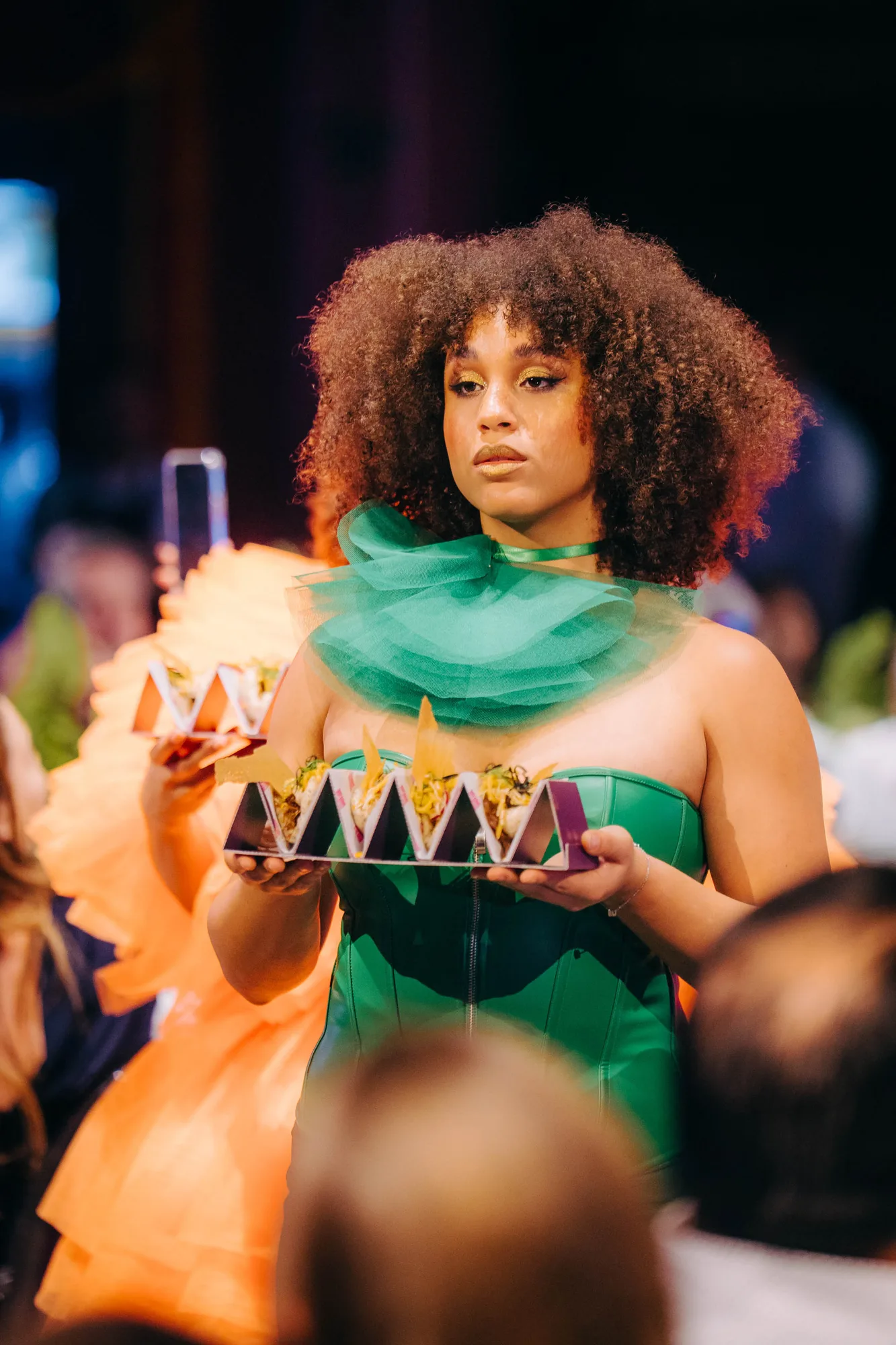 Woman with curly hair wearing a green dress and tulle collar, holding a tray of small food servings at an event.