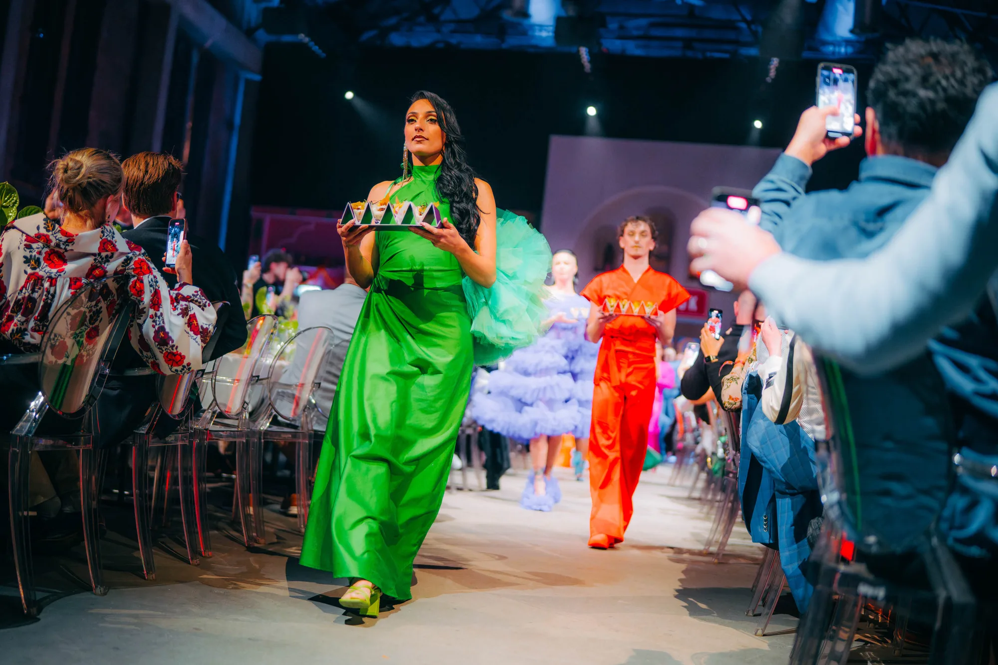 Models walking down the runway in colorful dresses holding trays with food during a fashion event.