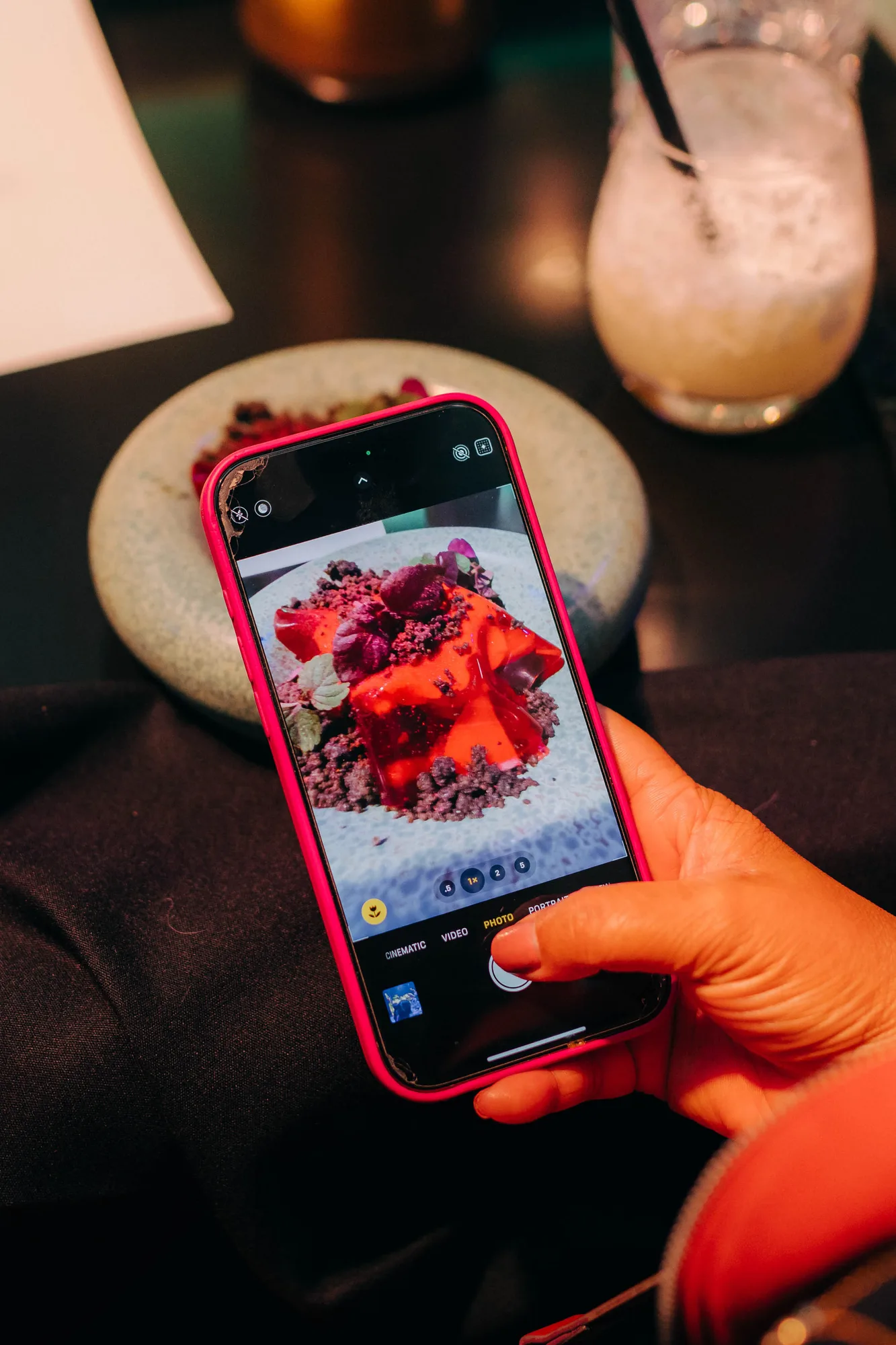 Hand holding a smartphone with a red case taking a photo of a dessert with red jelly and chocolate crumbs on a gray plate.
